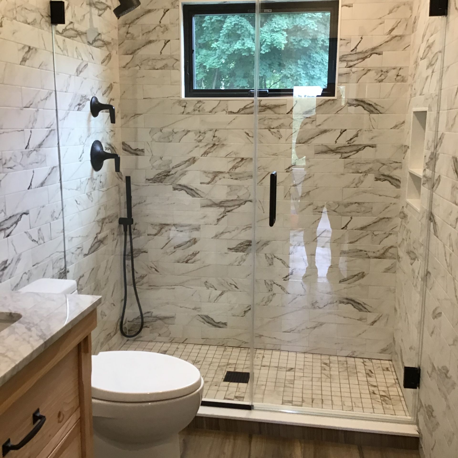 Modern marble-tiled bathroom with glass shower, white toilet, and small window above the shower.