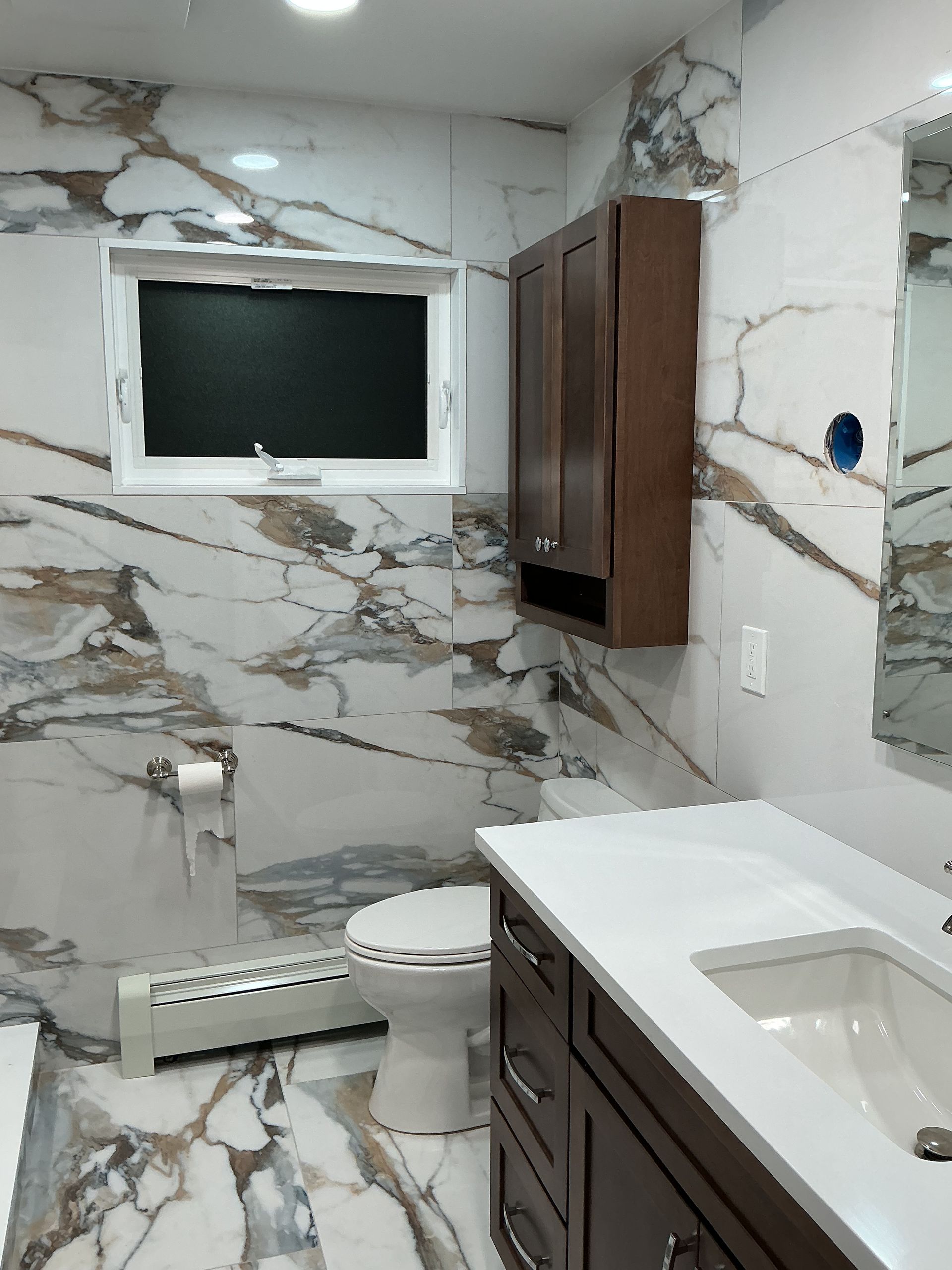 Modern bathroom with marble-pattern tile, dark wood cabinets, white sink, toilet, and a window.
