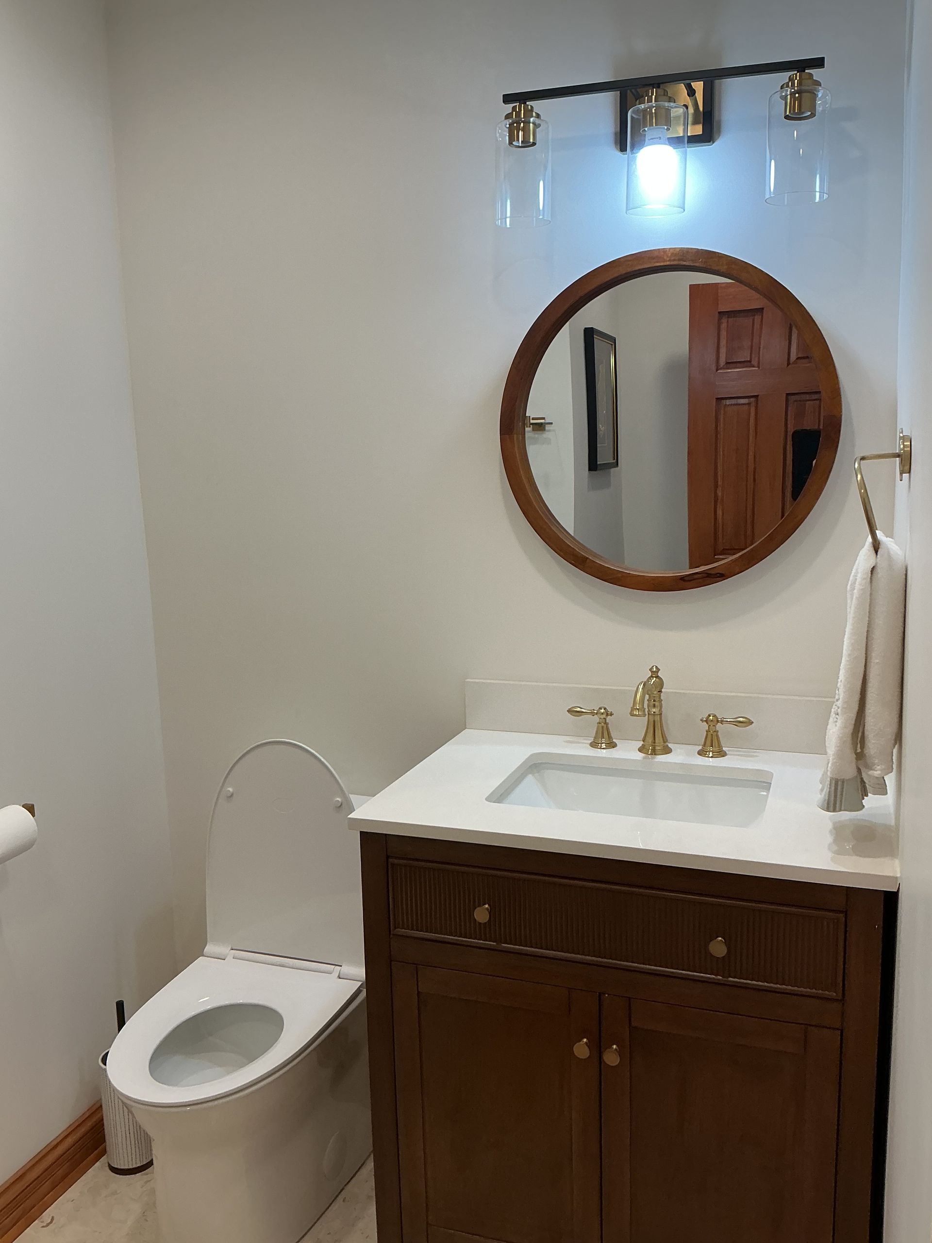 Small bathroom with a toilet, dark wood vanity, white sink, and round mirror.