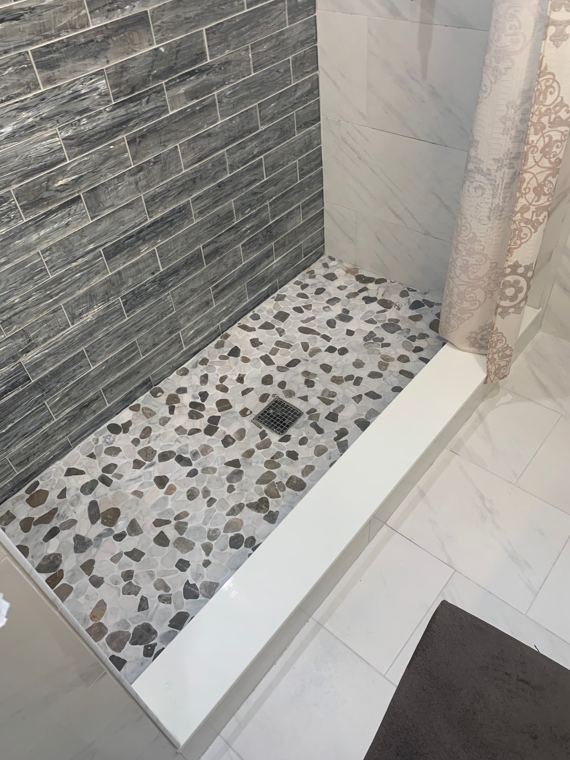 Empty shower stall with terrazzo floor, gray tile wall, and white curb next to a curtain.
