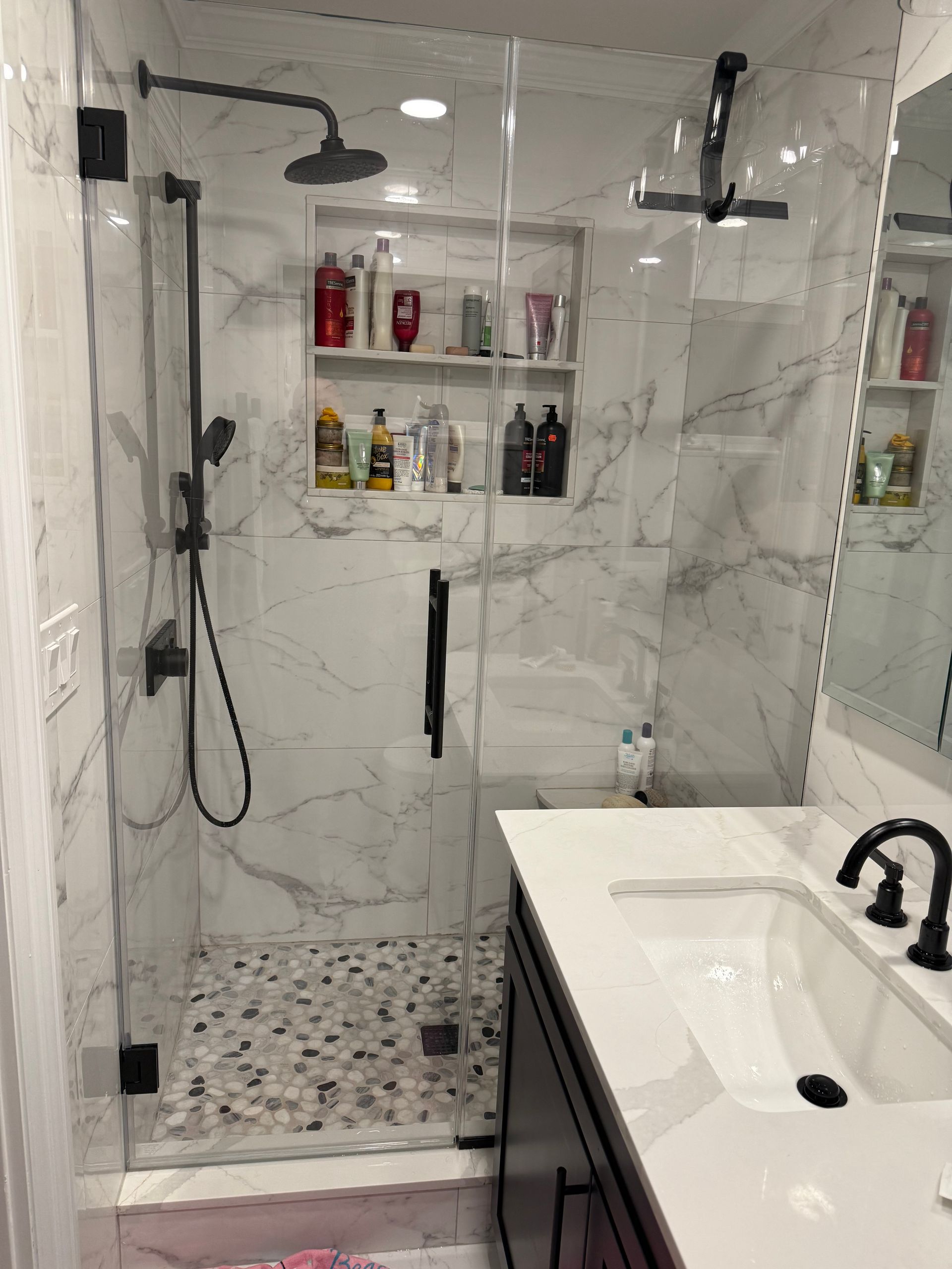 Modern marble-tiled bathroom with glass shower, built-in shelves, and white vanity sink.