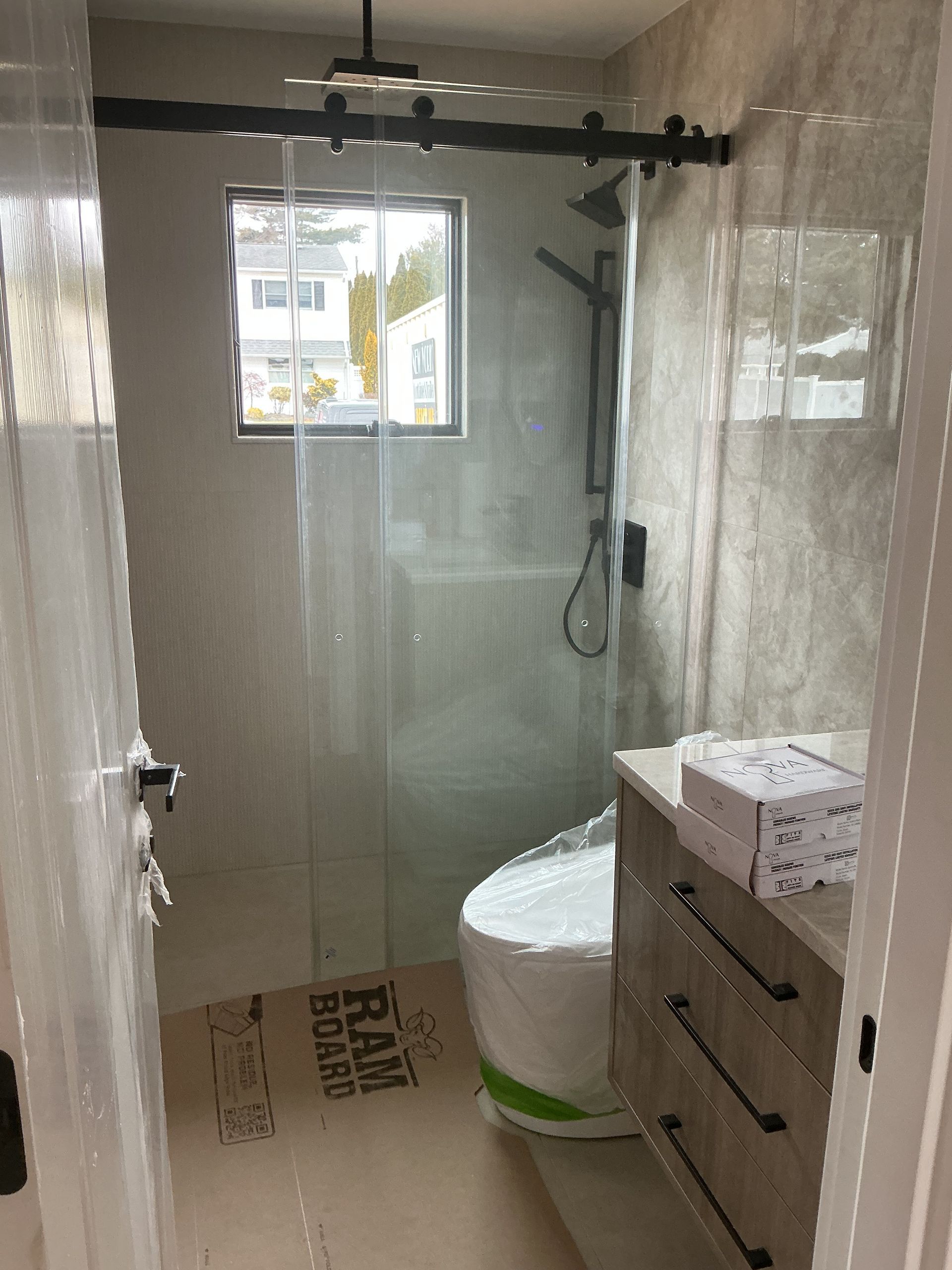 Small bathroom renovation with glass shower walls, window, toilet, and vanity under construction.