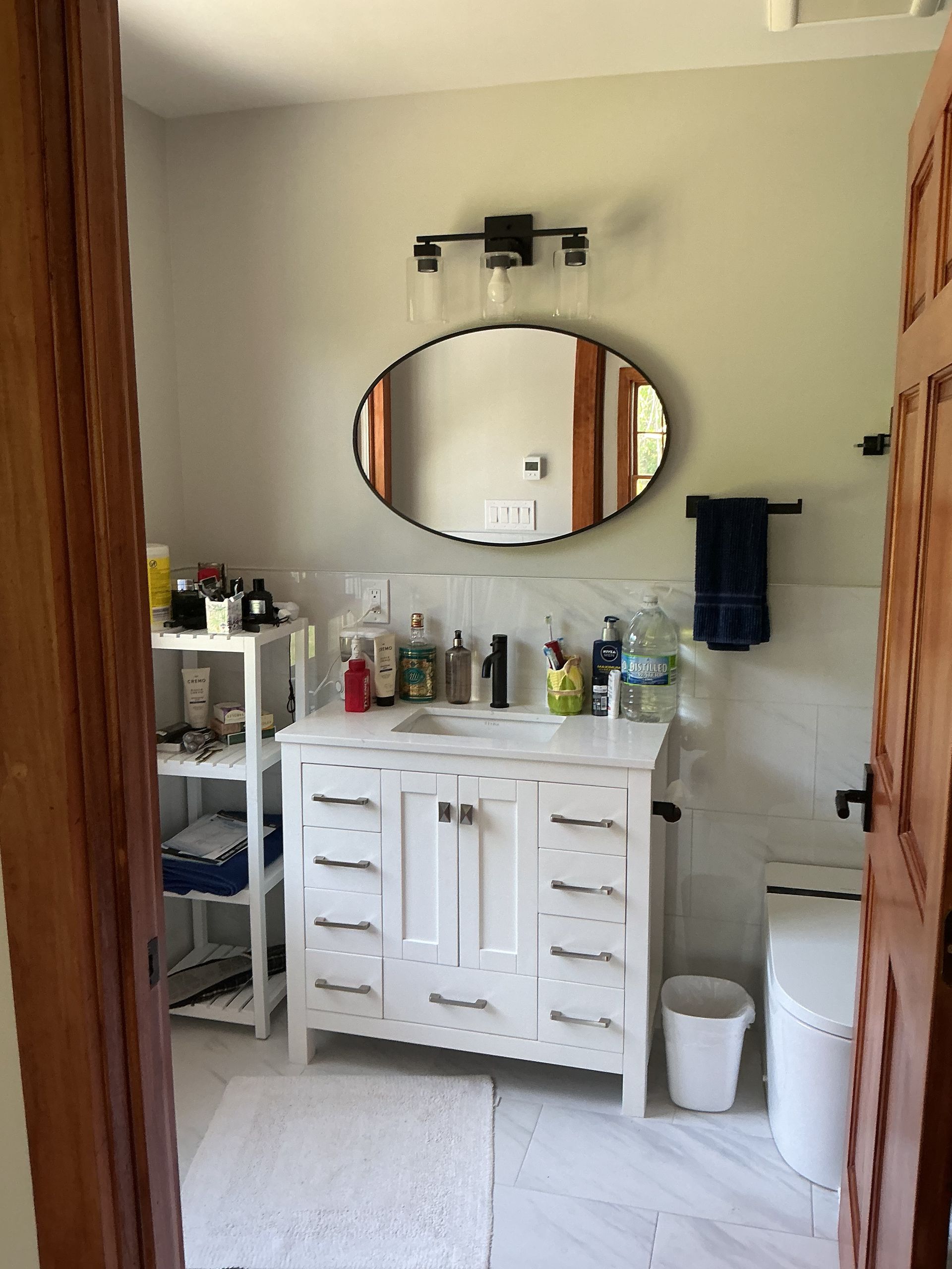 Small bathroom vanity with oval mirror, white cabinet, and shelves holding toiletries.