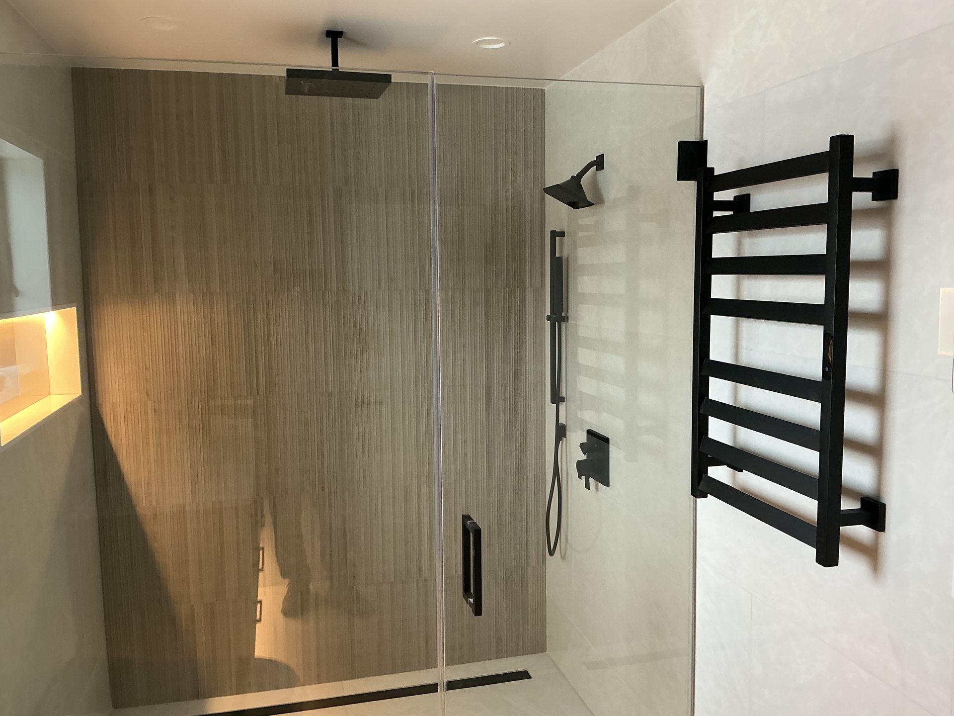 Modern shower with glass door, beige tile walls, black towel rack, and showerhead.