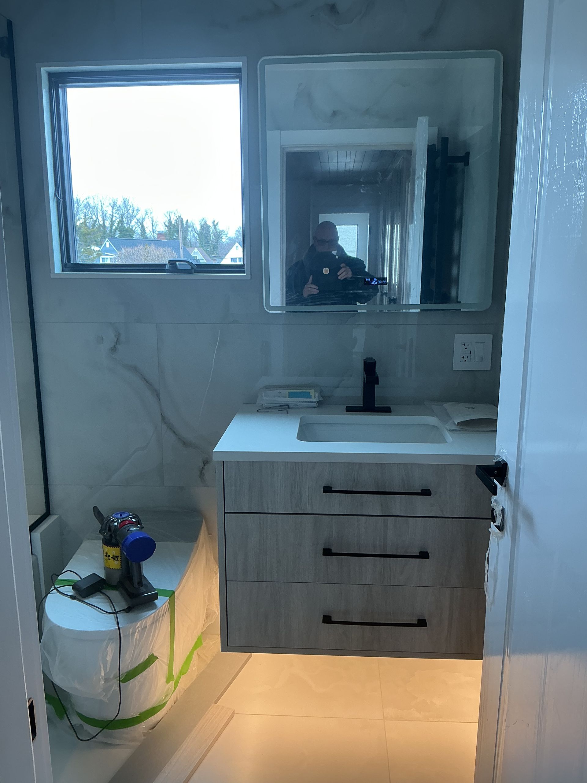 Small bathroom with vanity, mirror, window, exposed wiring, and a bucket on the floor under warm light