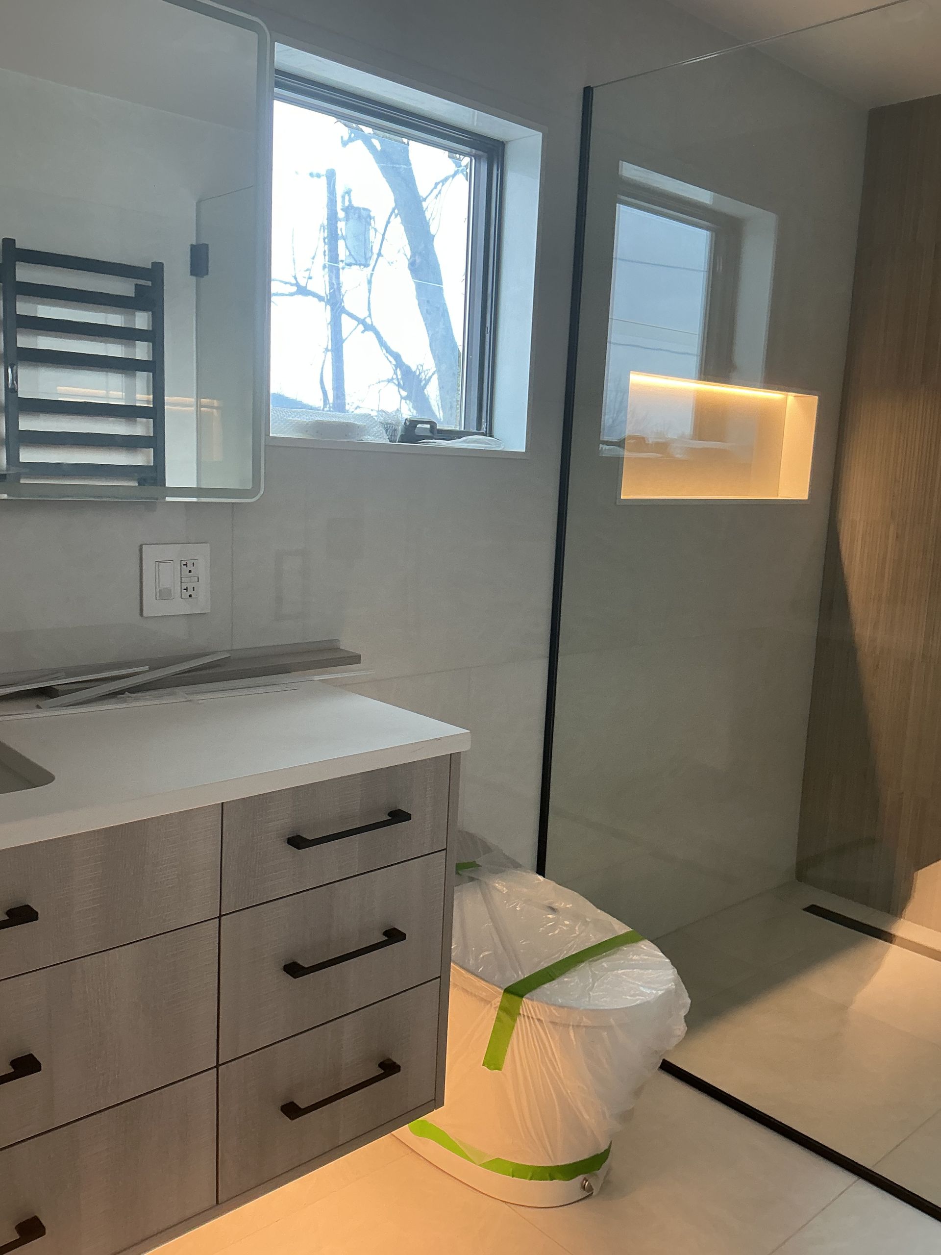 Bathroom vanity with gray drawers, lit mirror, and a wrapped box on the floor near a glass shower.