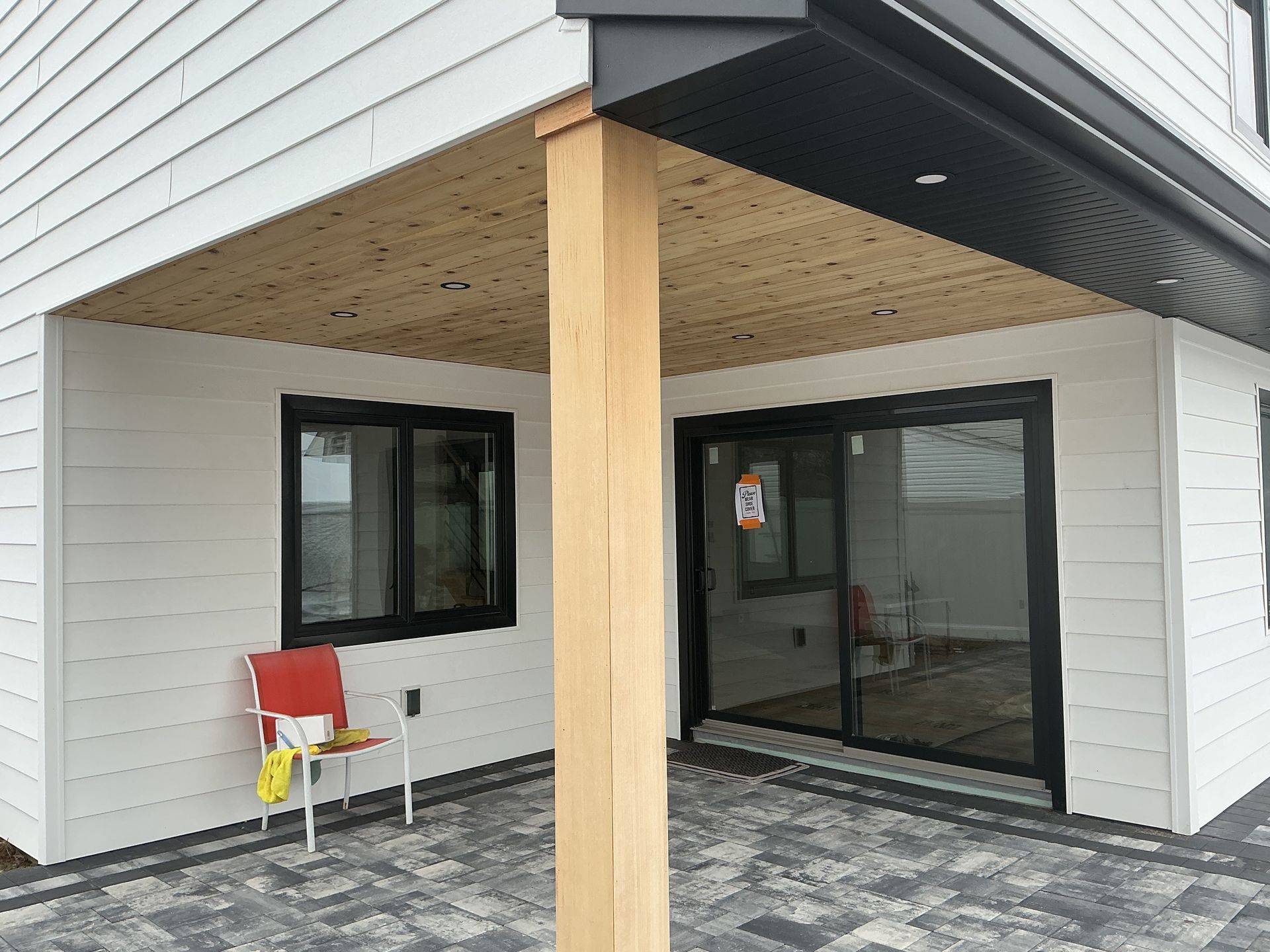 Covered porch entrance with wooden post, white siding, black windows, and a red chair on the left.