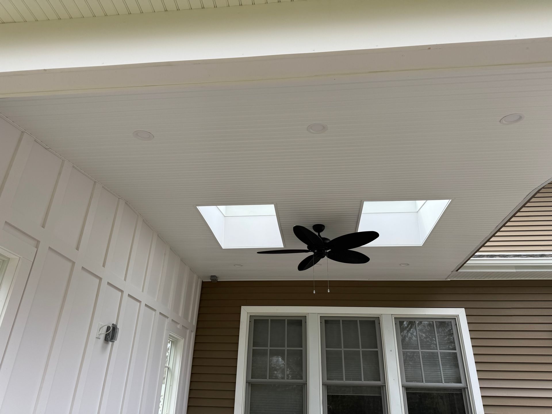 Covered porch ceiling with two skylights and a black ceiling fan above a window and siding