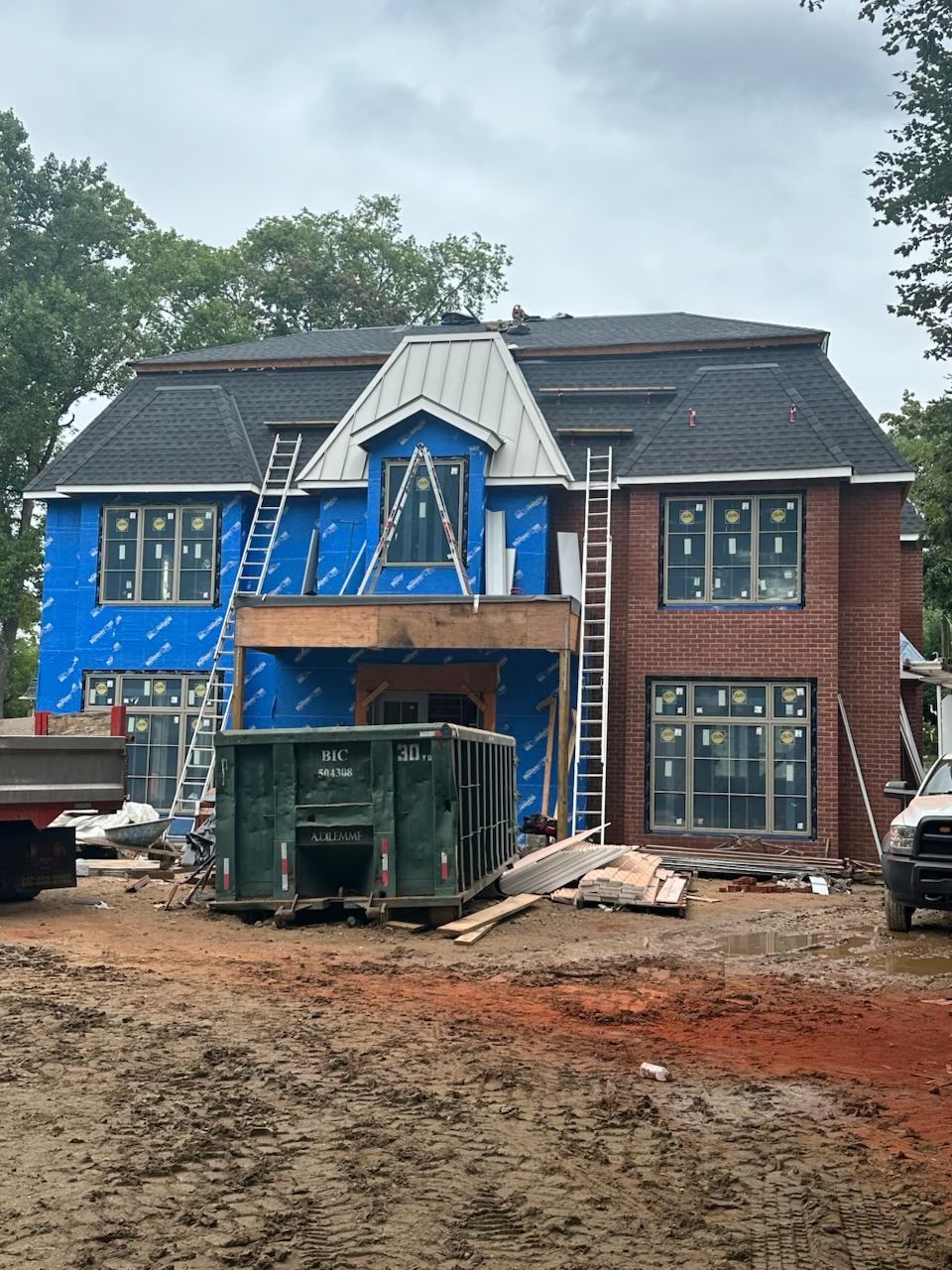 Two-story house under construction with blue wrap, scaffolding, and dumpsters in front