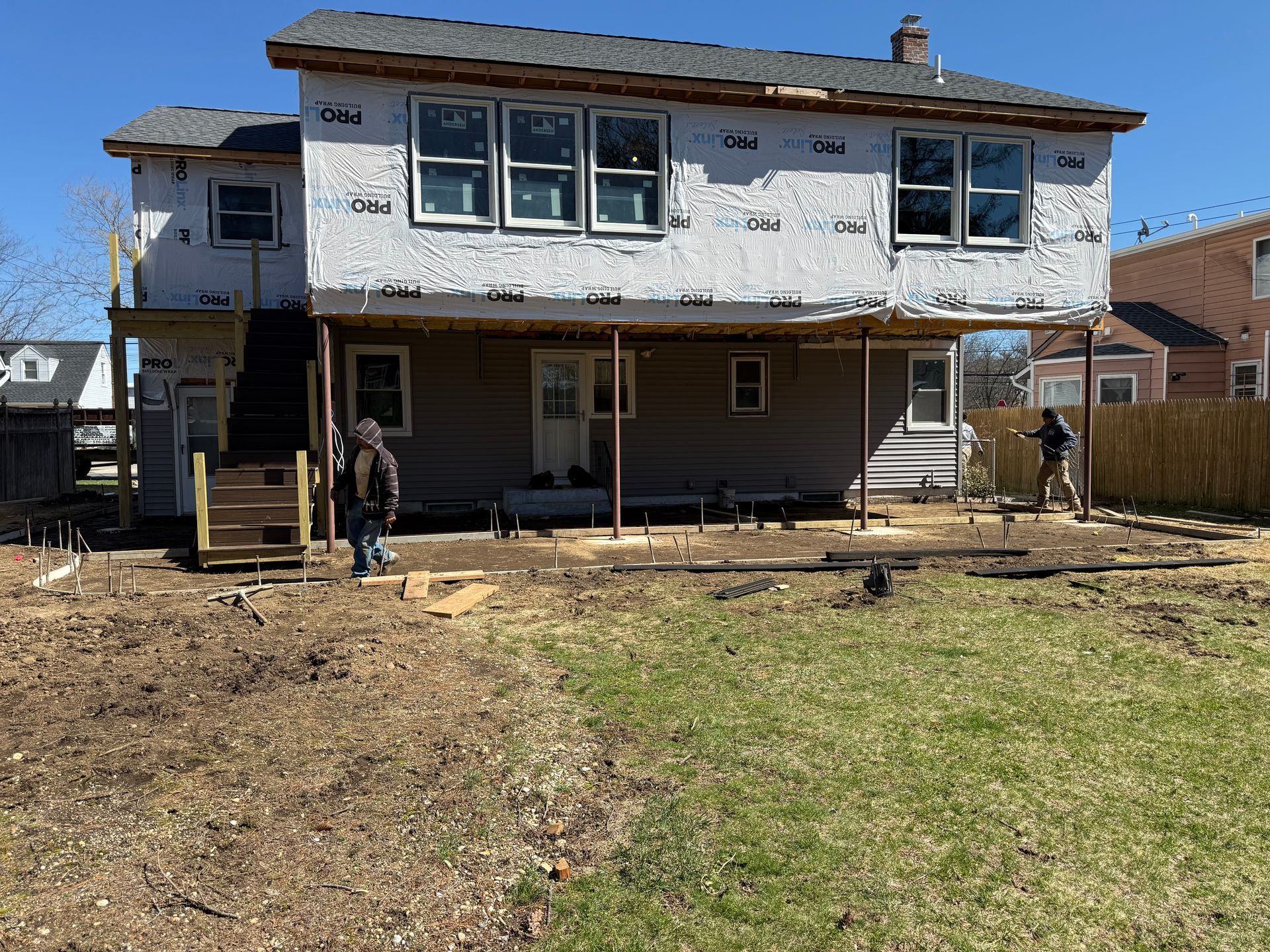 Backyard view of a two-story house under renovation with workers on the porch and bare ground