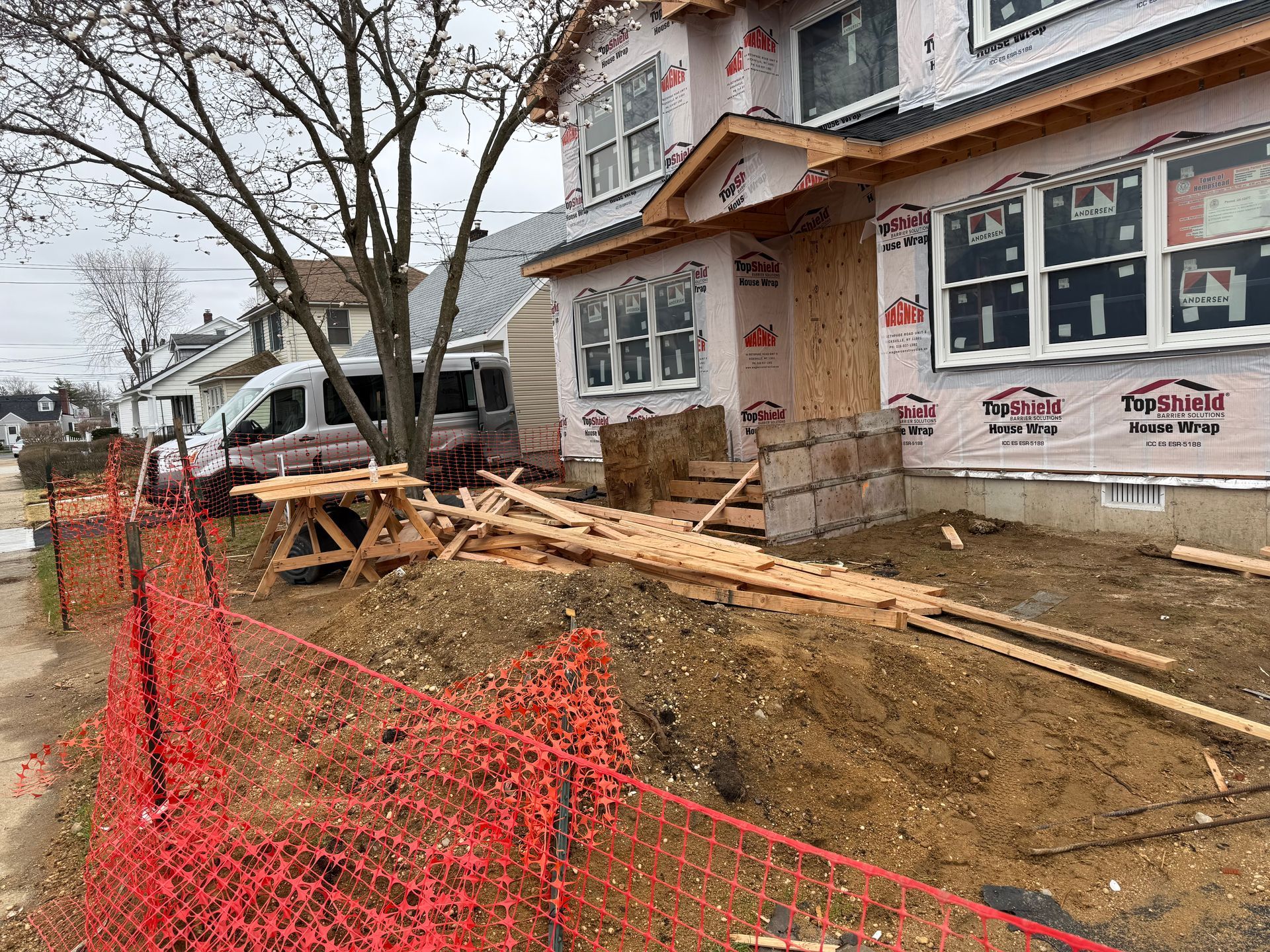 Construction site outside a house with dirt trench, orange safety fencing, and lumber stacked near the porch.