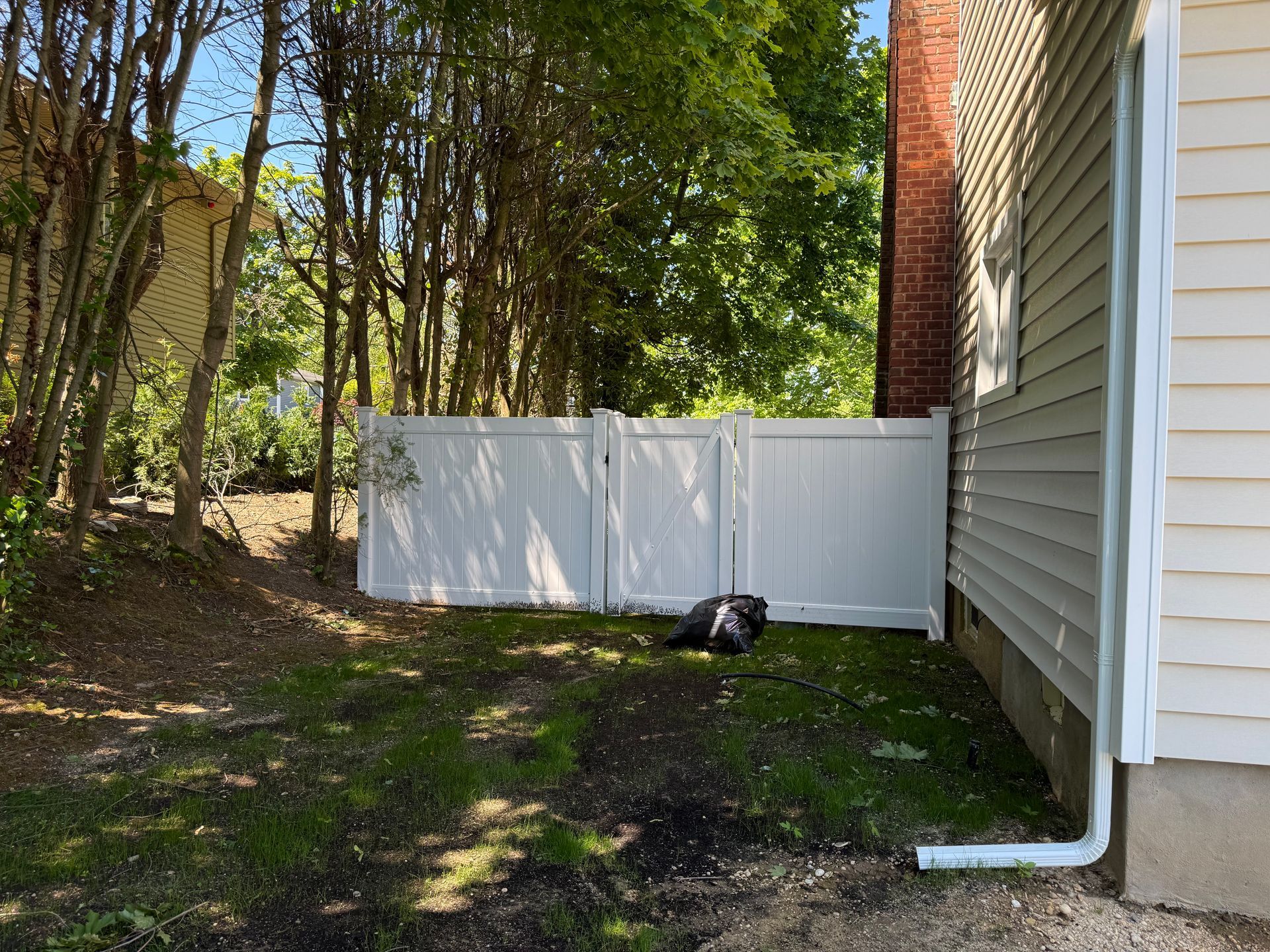 Backyard beside a house with a white fence, trees, and a small black bag on the ground.