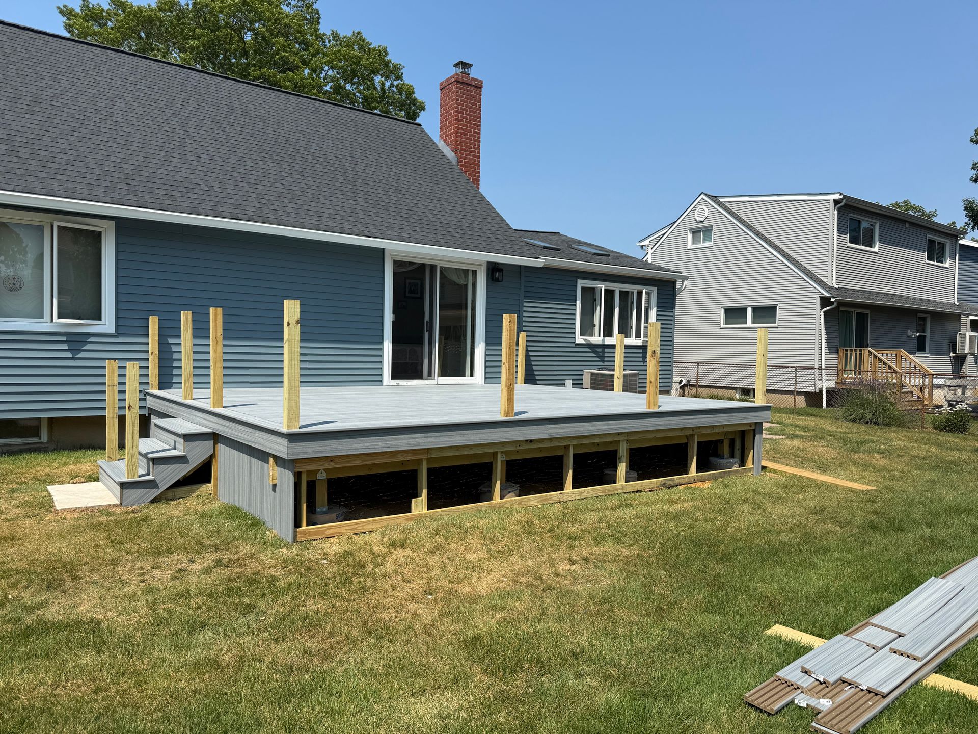 Blue house backyard with a raised deck under construction and unfinished posts on a grassy lawn