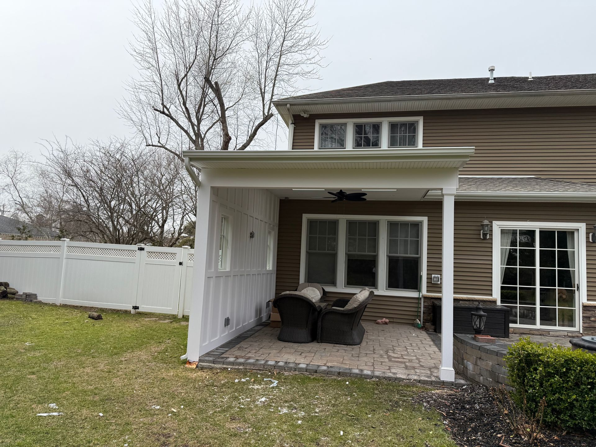Covered patio with white columns, brown siding, wicker chairs, and a fenced yard.