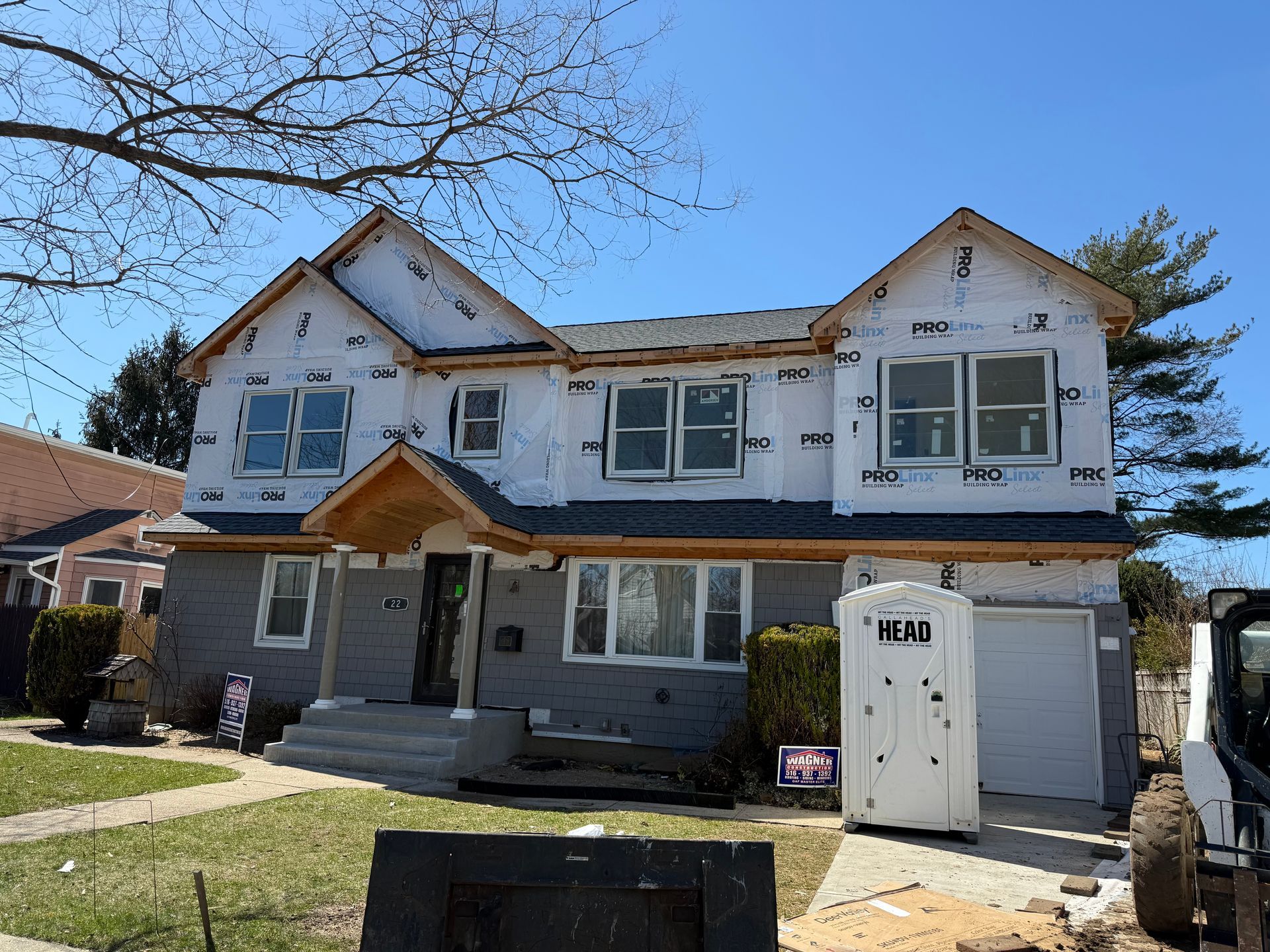 Two-story house under construction with white siding, exposed roof framing, and a portable toilet in the driveway