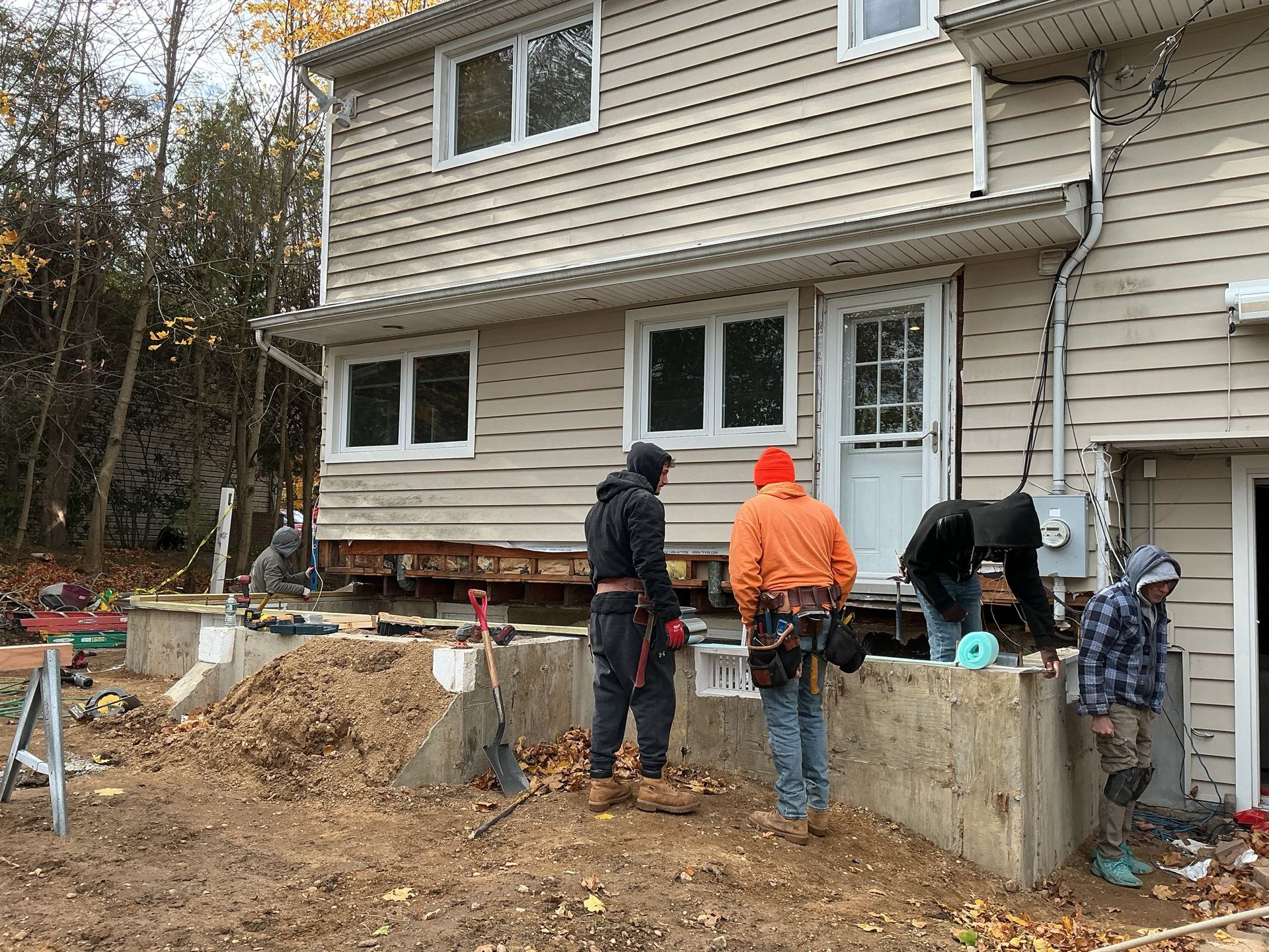 Workers building a raised concrete patio beside a beige house, with lumber and dirt piled nearby