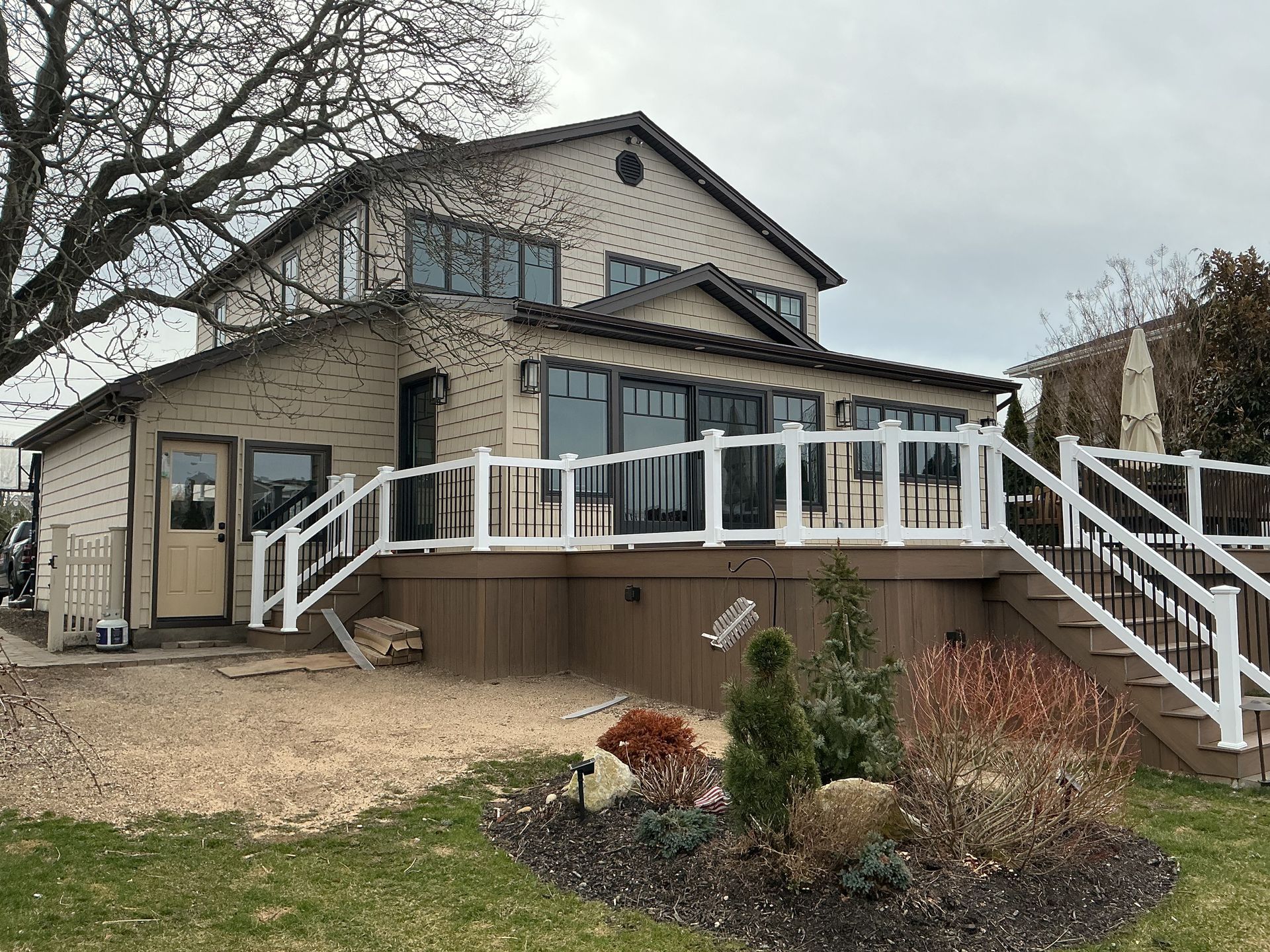 Two-story beige house with white deck railings and stairs, a small front garden, and leafless trees