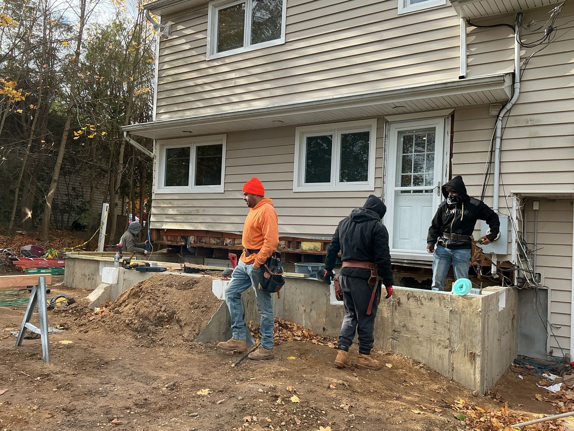 Workers repairing a house foundation beside a dirt-covered yard and concrete wall