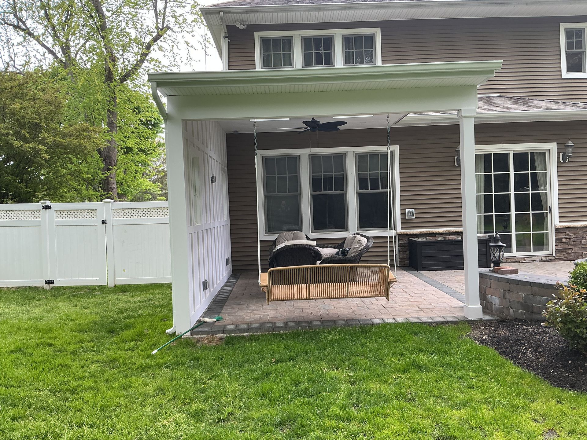 Covered patio with white columns, black sofa, wicker table, and white fence beside a house.