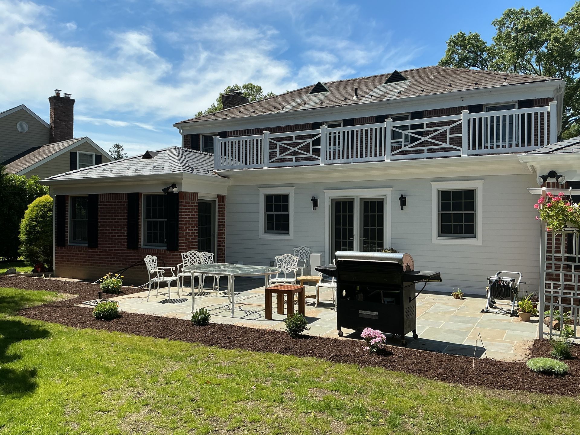 Back patio with white house, brick grill, patio table, chairs, and green lawn under a blue sky