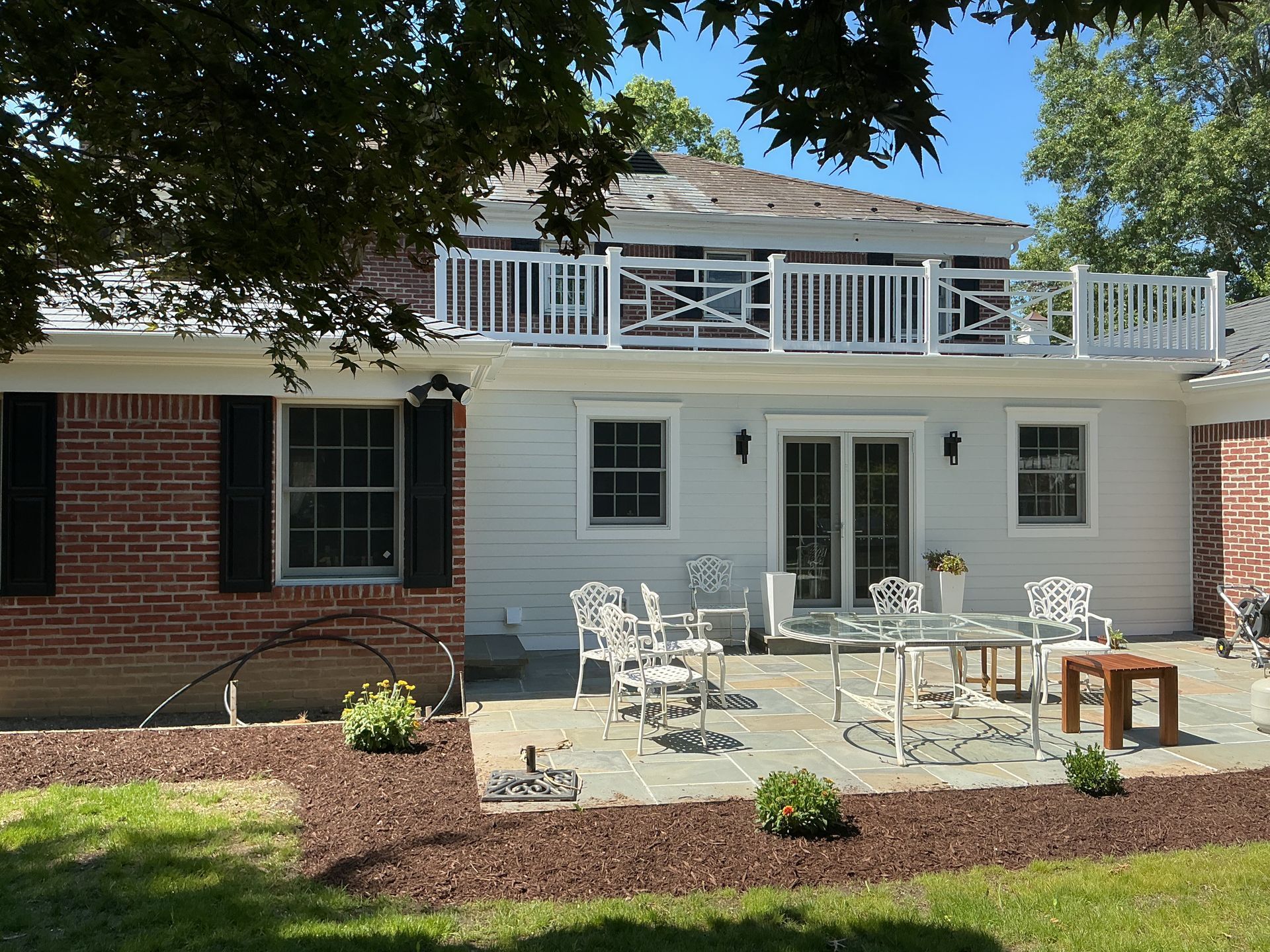 Patio with white outdoor tables and chairs beside a brick-and-white house under trees