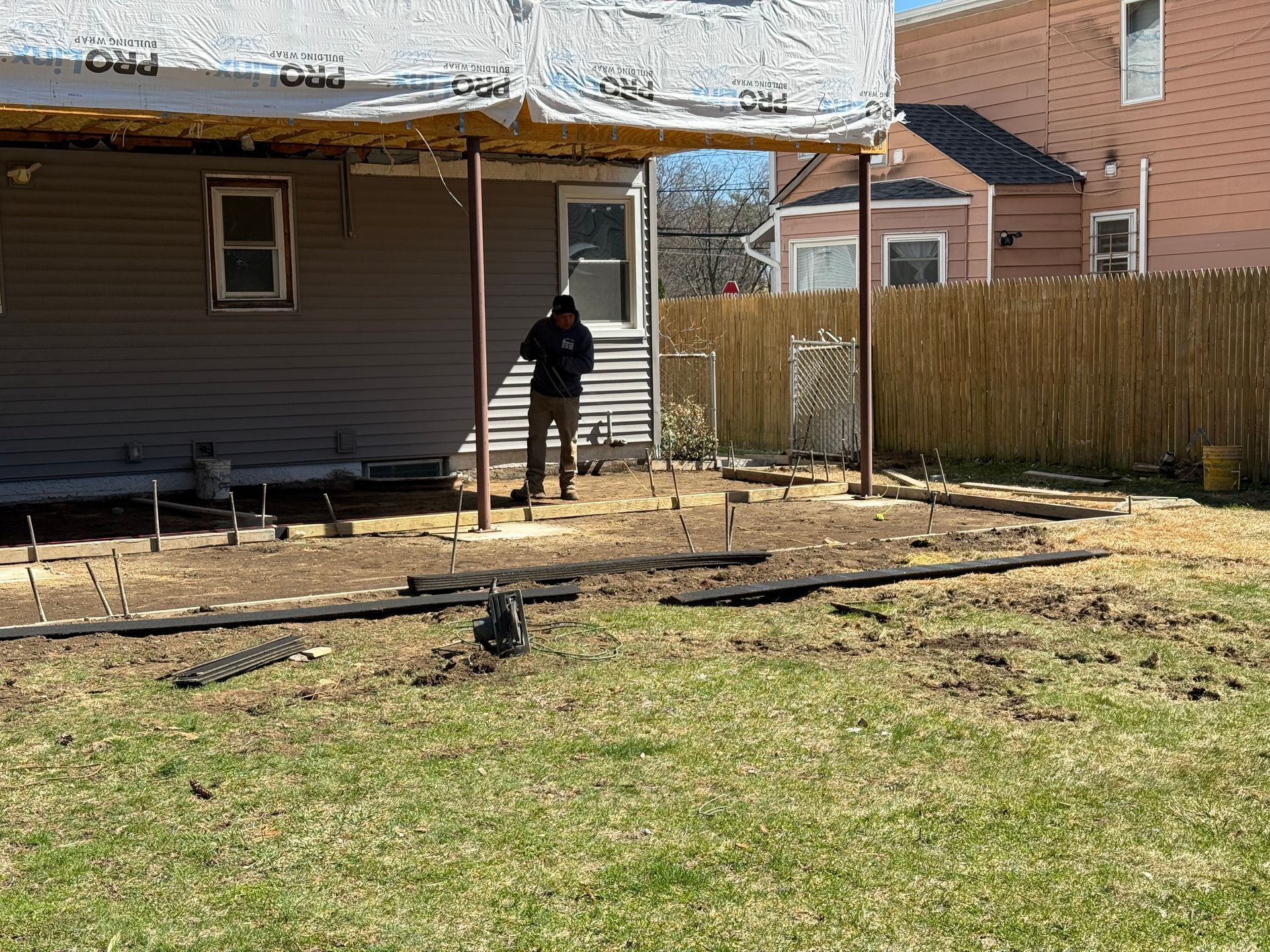 Person standing in a backyard near a house, with a deck, fence, and bare ground.
