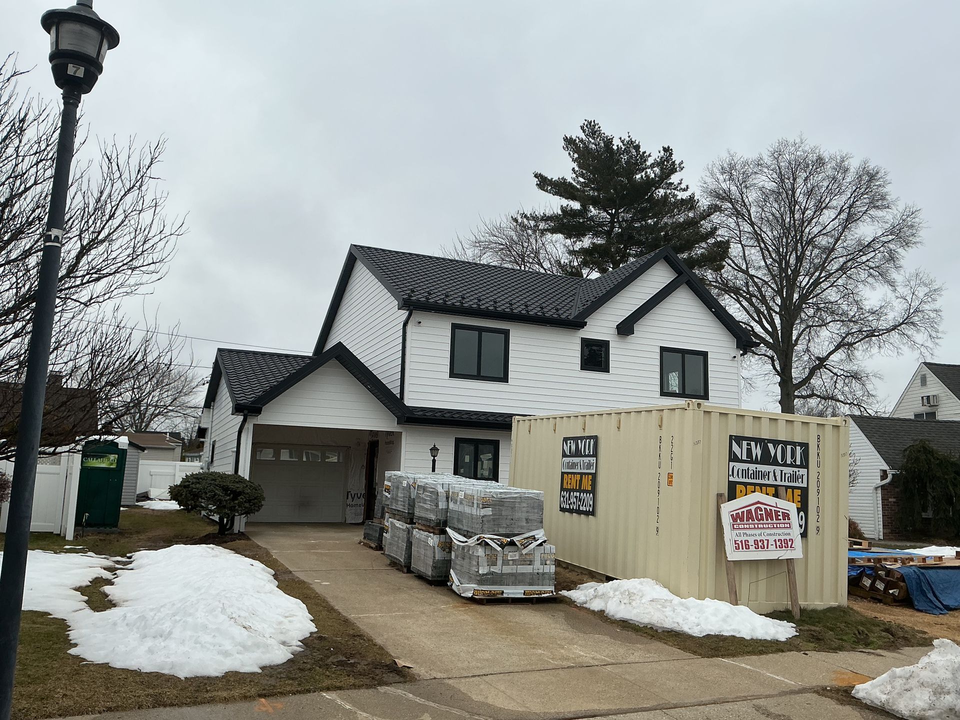 House under renovation with stacked building materials, snow patches, and a beige construction fence in front