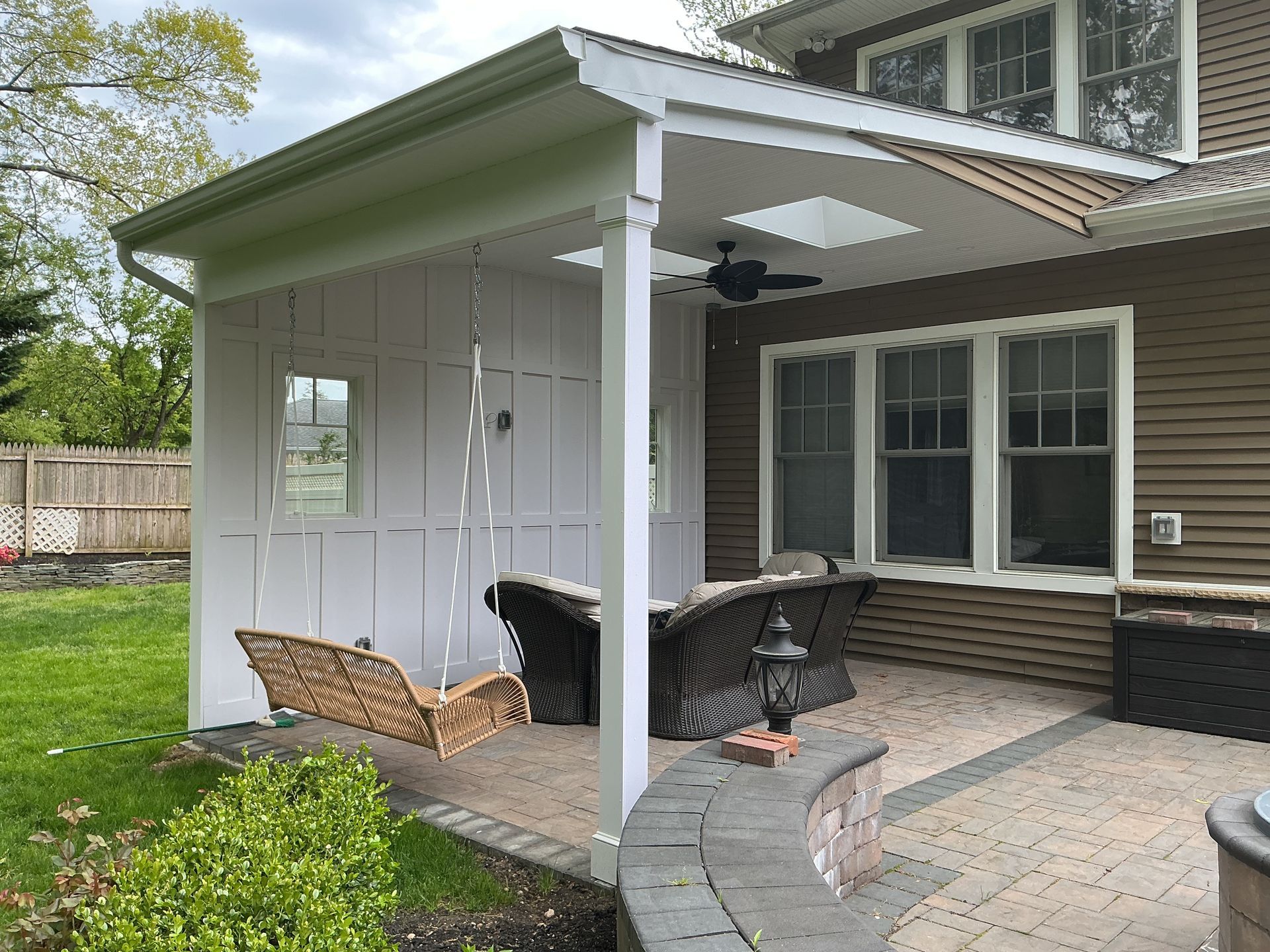 Covered patio with ceiling fan, wicker chairs, and a grill beside a house with white siding.