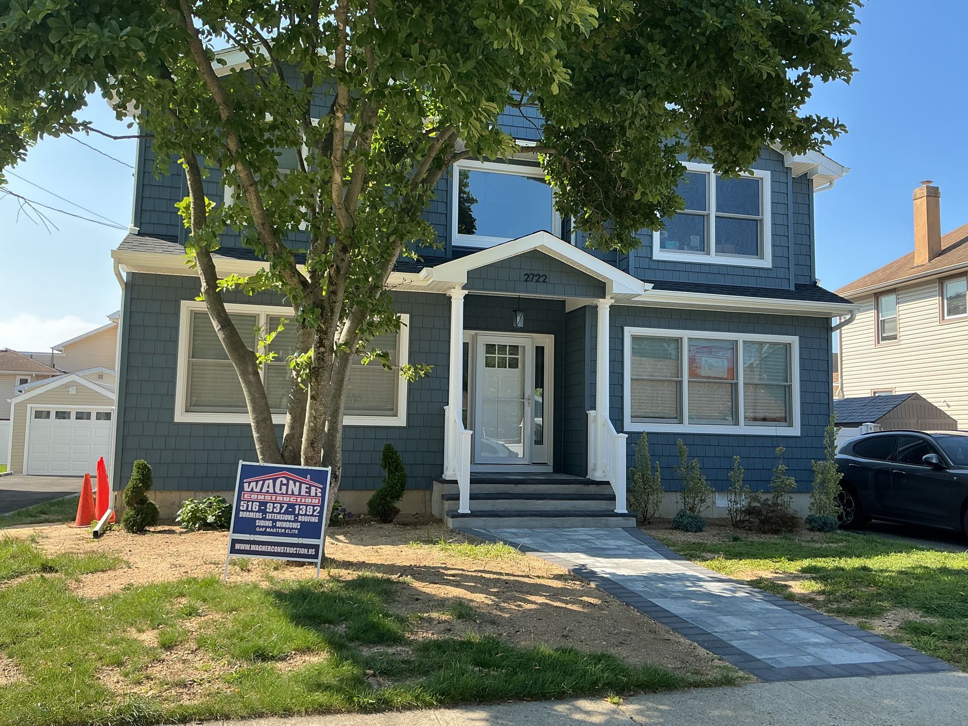 Blue two-story house with a front porch and a real estate sign in the yard, shaded by a large tree.