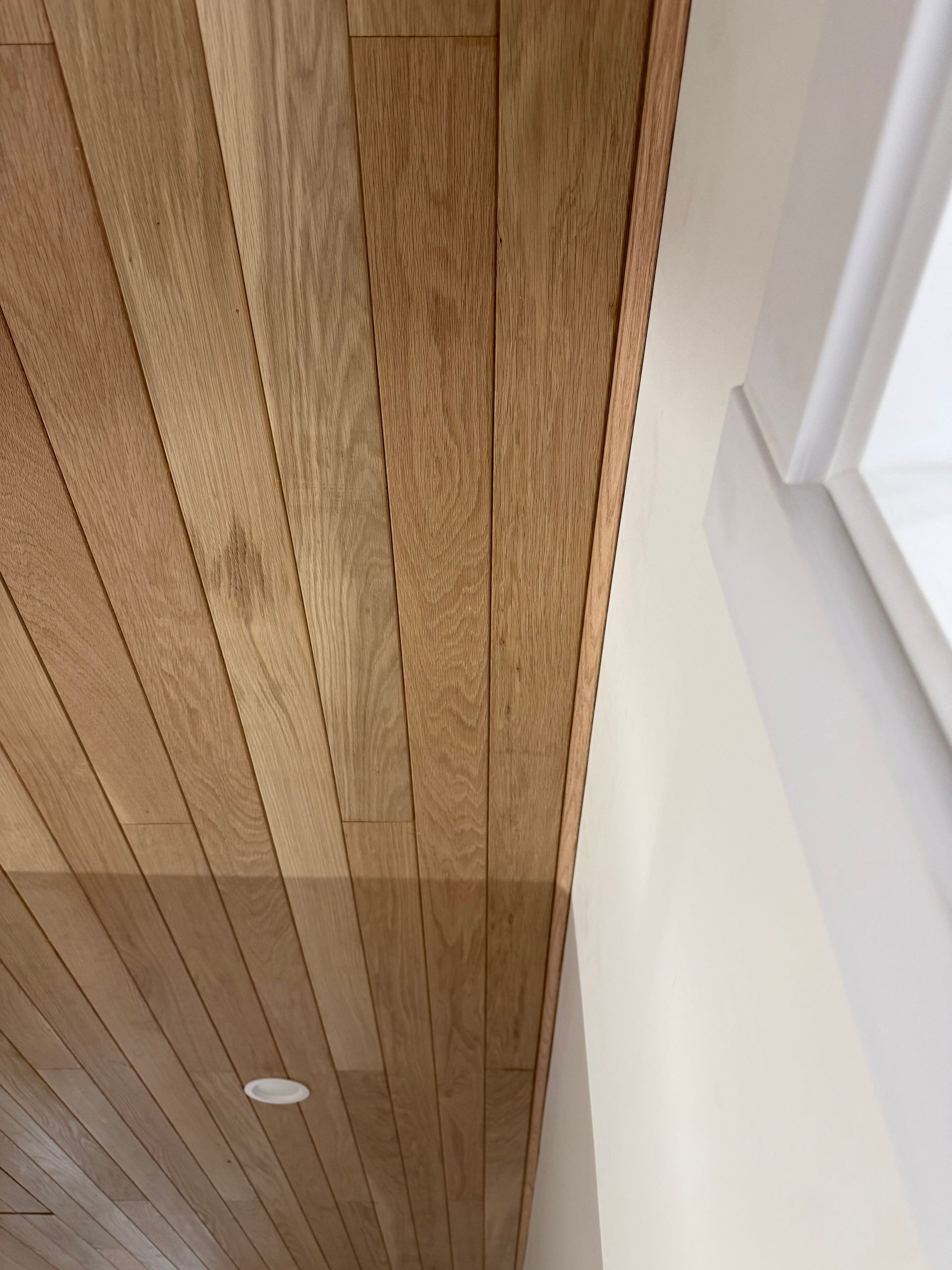 Wooden slatted ceiling beside a white wall with a window frame and recessed light.