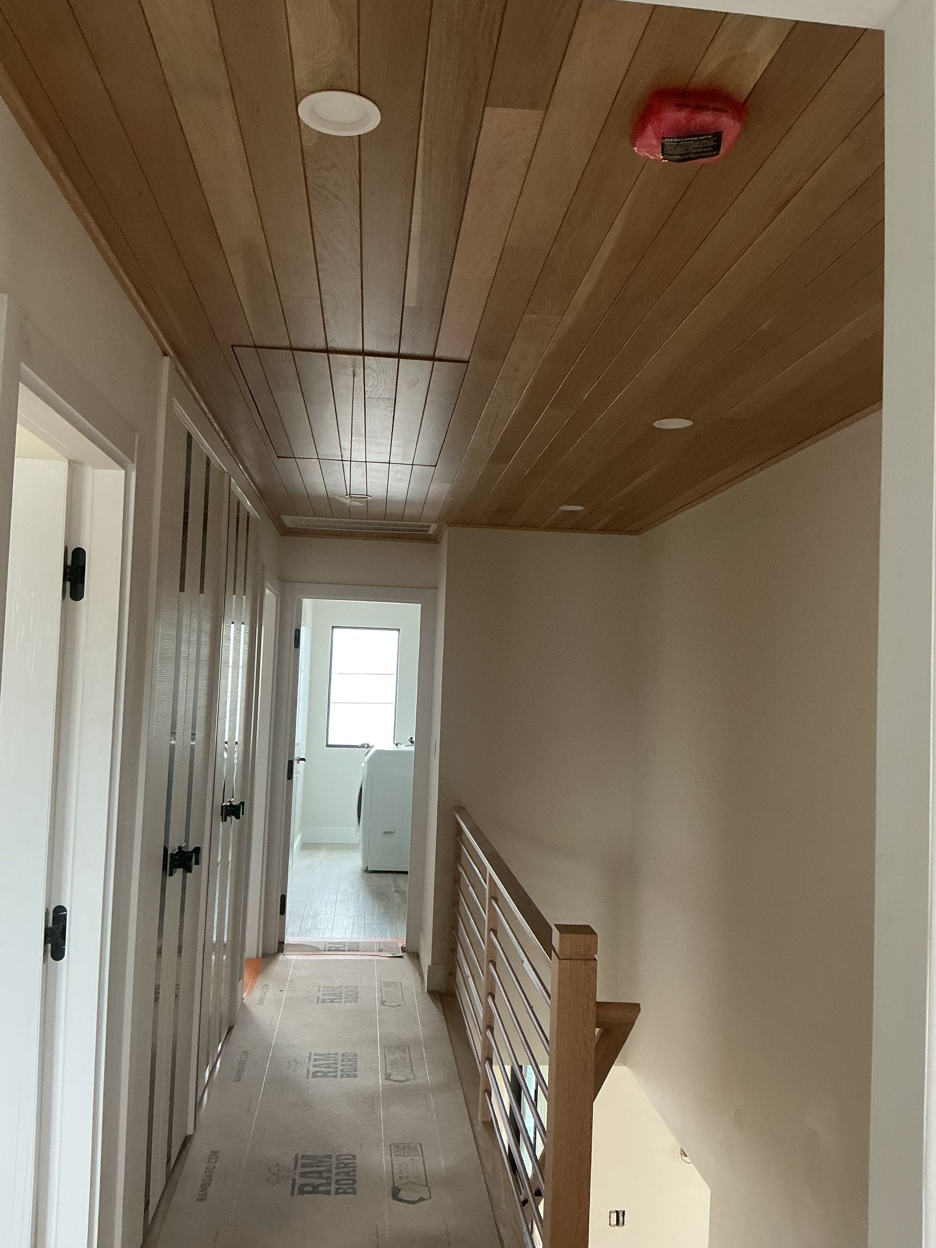 Narrow hallway with wood ceiling, white walls, and a railing leading to a bright room.
