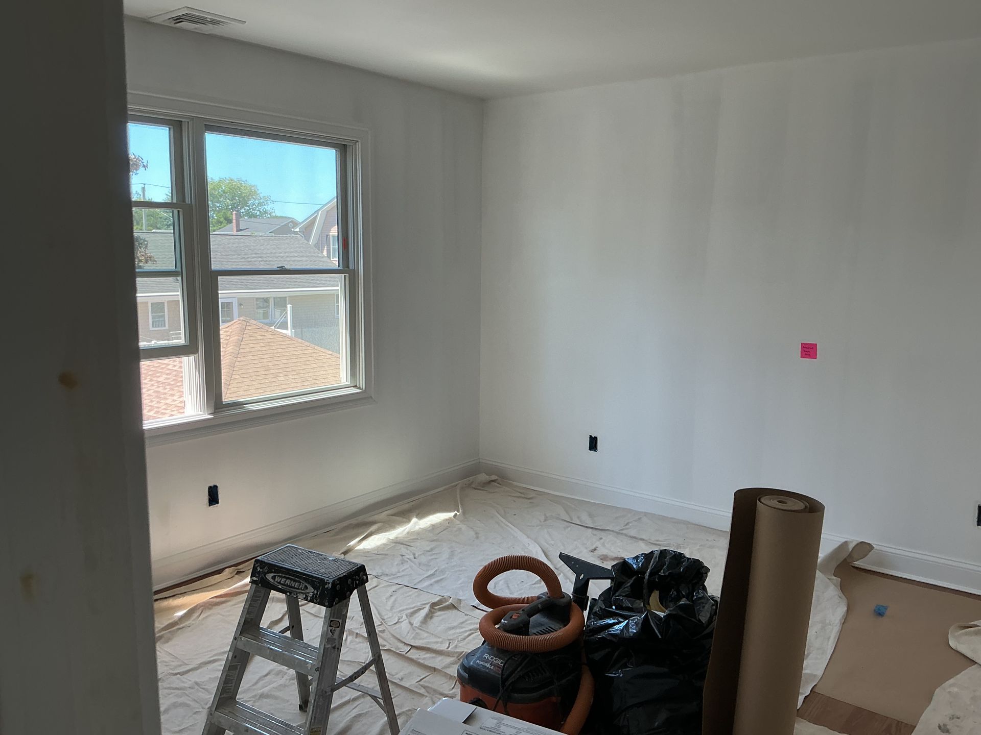Empty room under renovation with white walls, window light, a step ladder, and construction materials on the floor