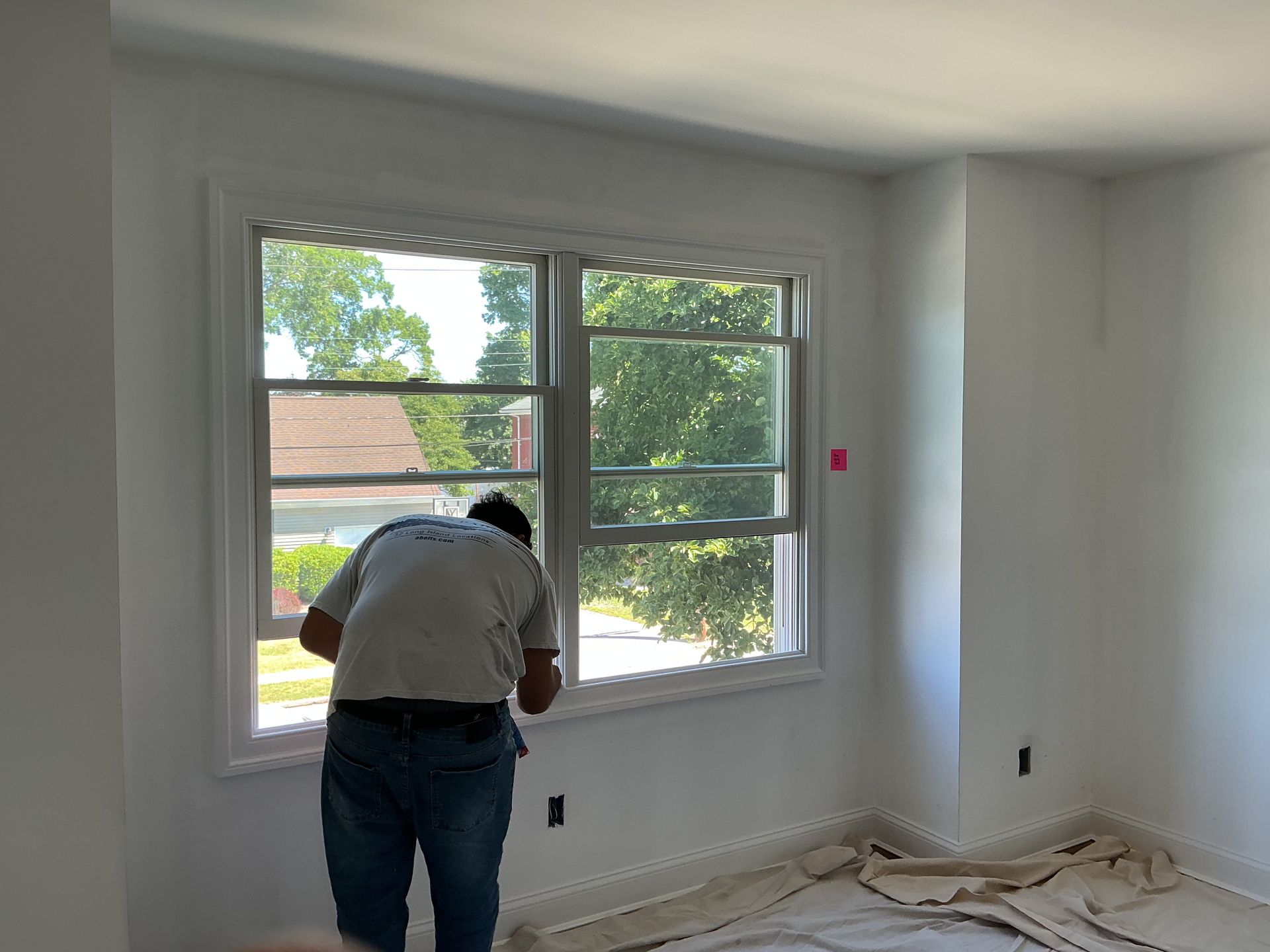 Person painting white window trim in a bright empty room with a large window and greenery outside