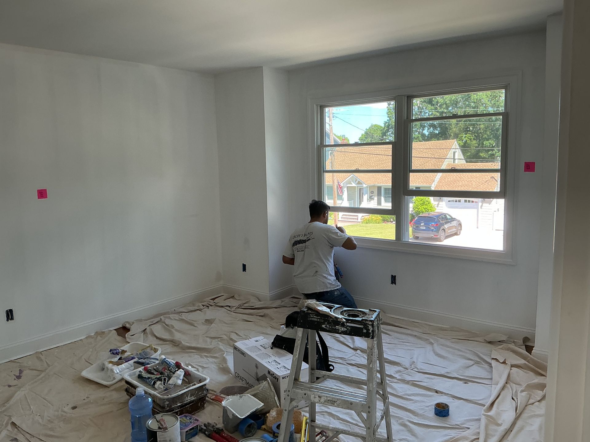 Person painting a white room near a window, with paint supplies and drop cloths on the floor.
