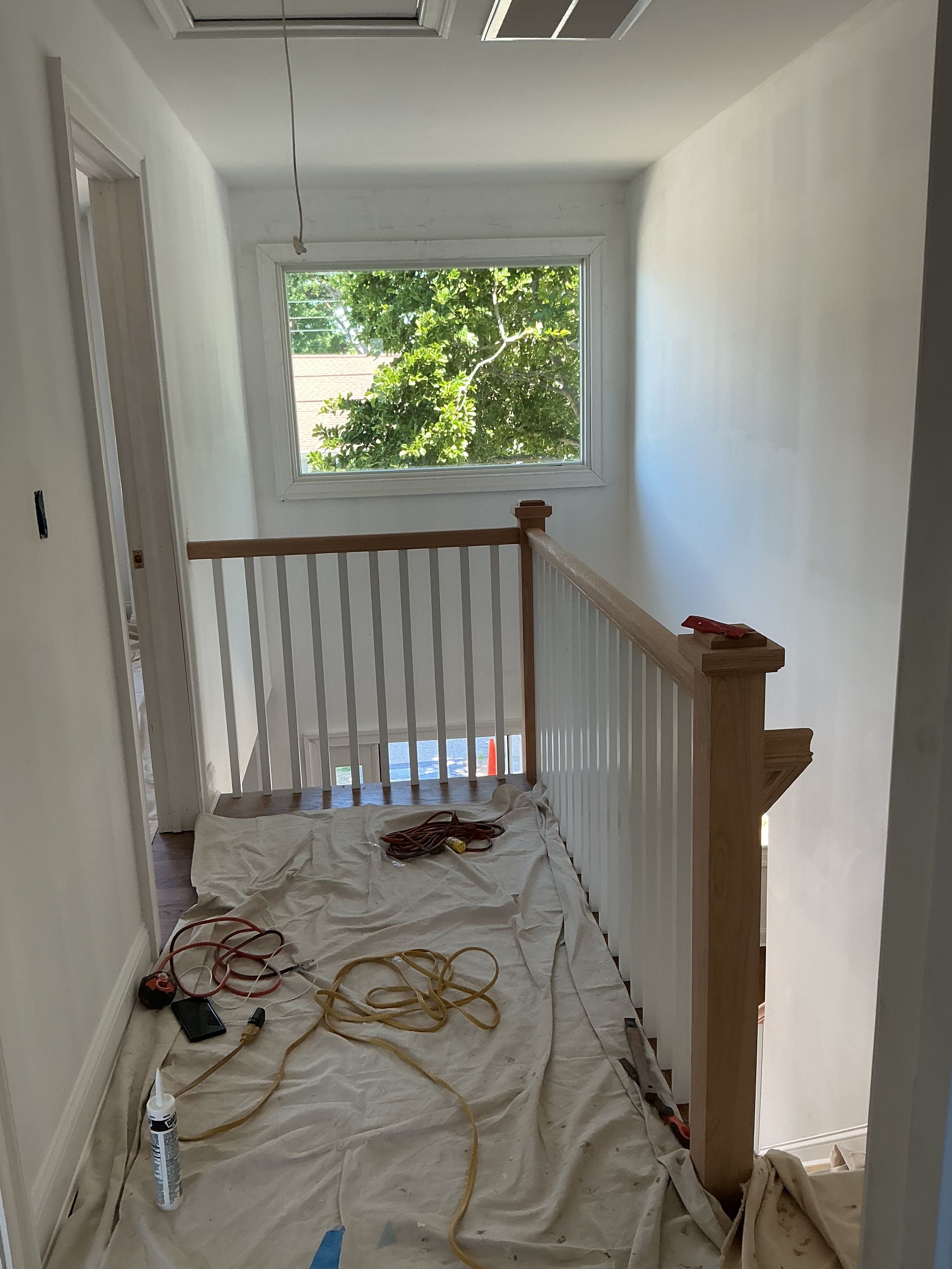 Hallway with a stair landing, white walls, wood railing, and drop cloths/tools on the floor