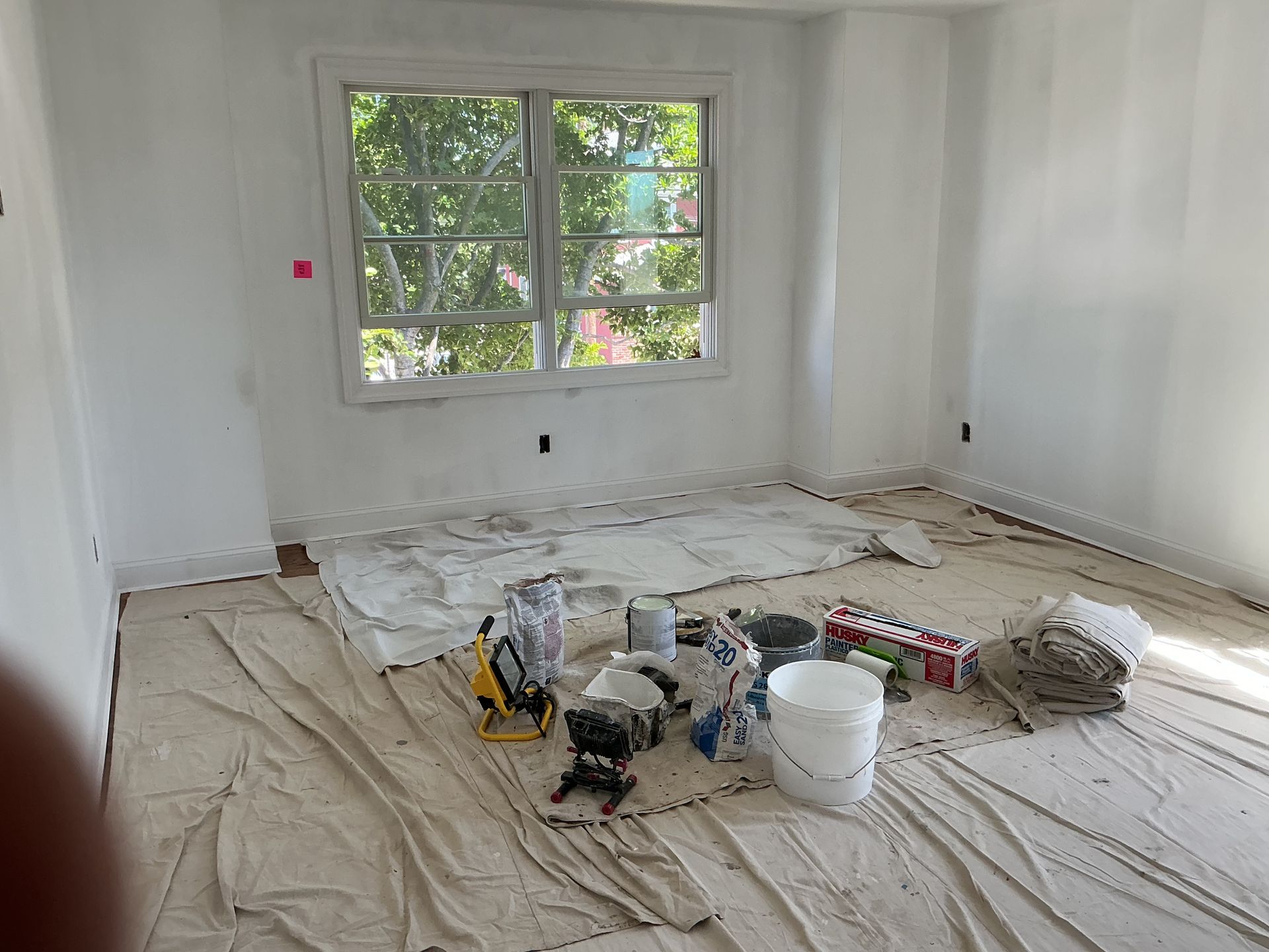 Renovation room with covered floor, paint supplies, and a large window letting in daylight