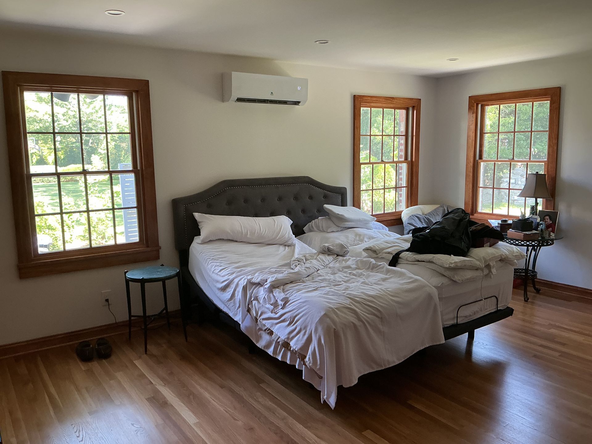 Bedroom with an unmade bed, white walls, wood floors, and windows letting in daylight.