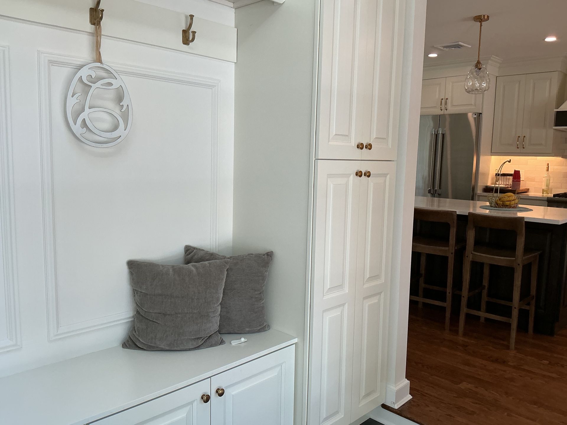 White entryway bench with gray pillows beside a doorway leading to a kitchen and wooden floor