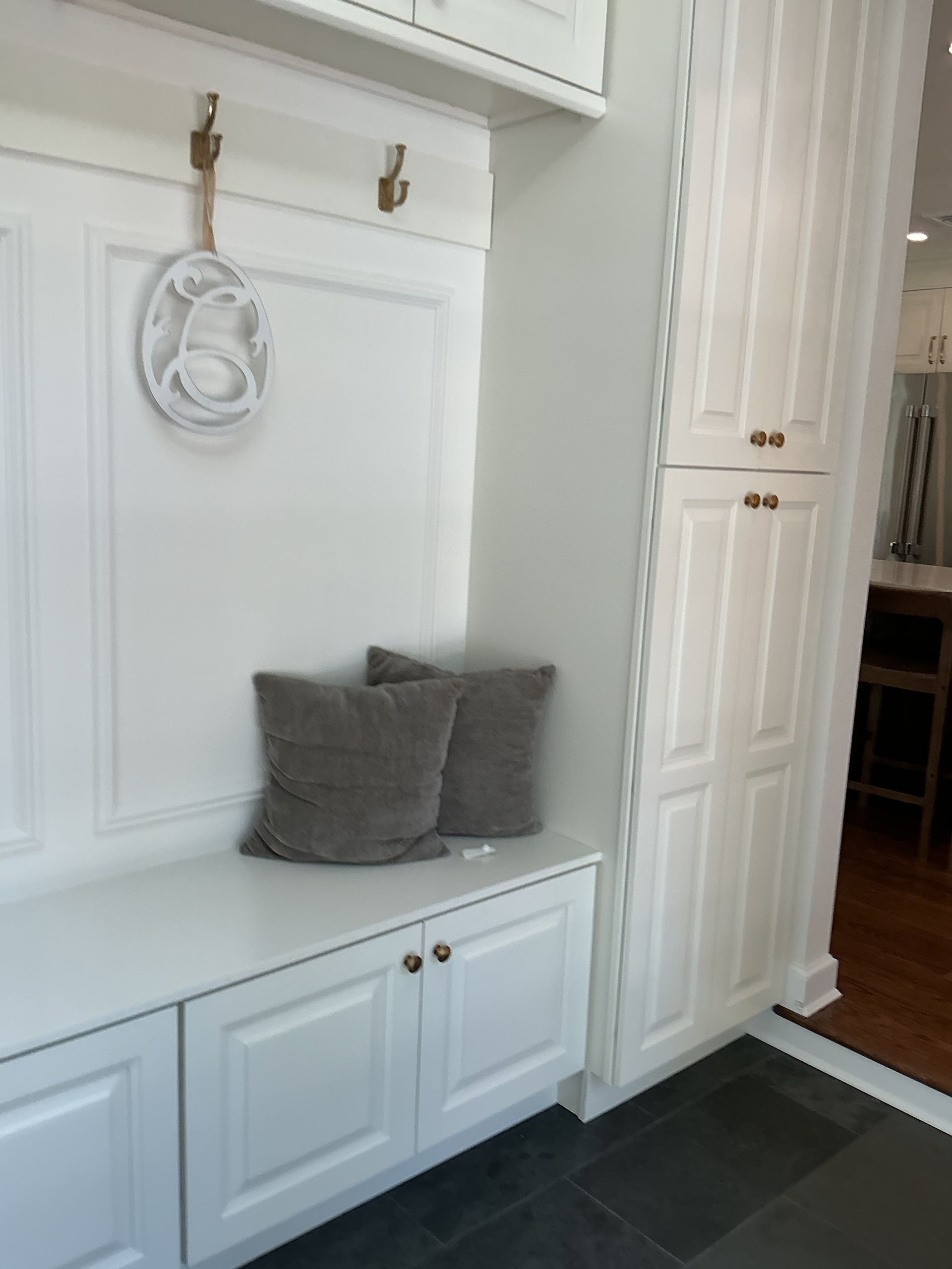 White entryway bench with gray cushions and hooks, beside a tall white cabinet and dark tile floor