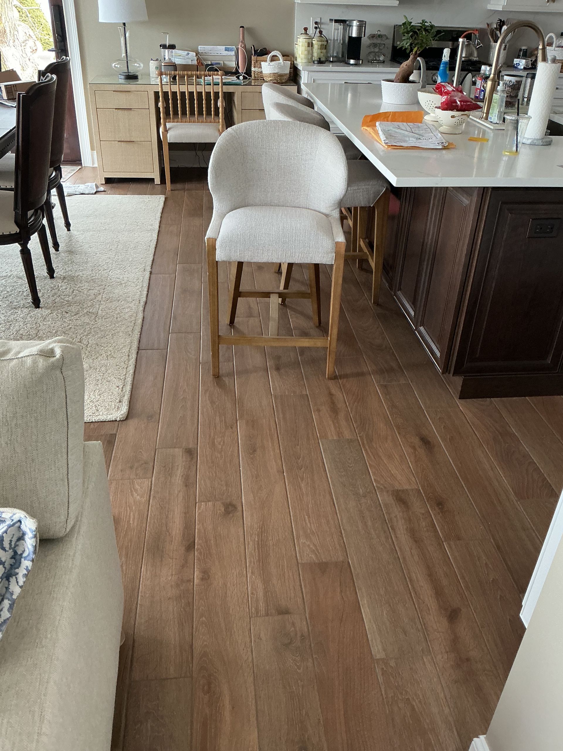 Kitchen with a white upholstered stool at a counter, hardwood floor, and an open dining area in the background