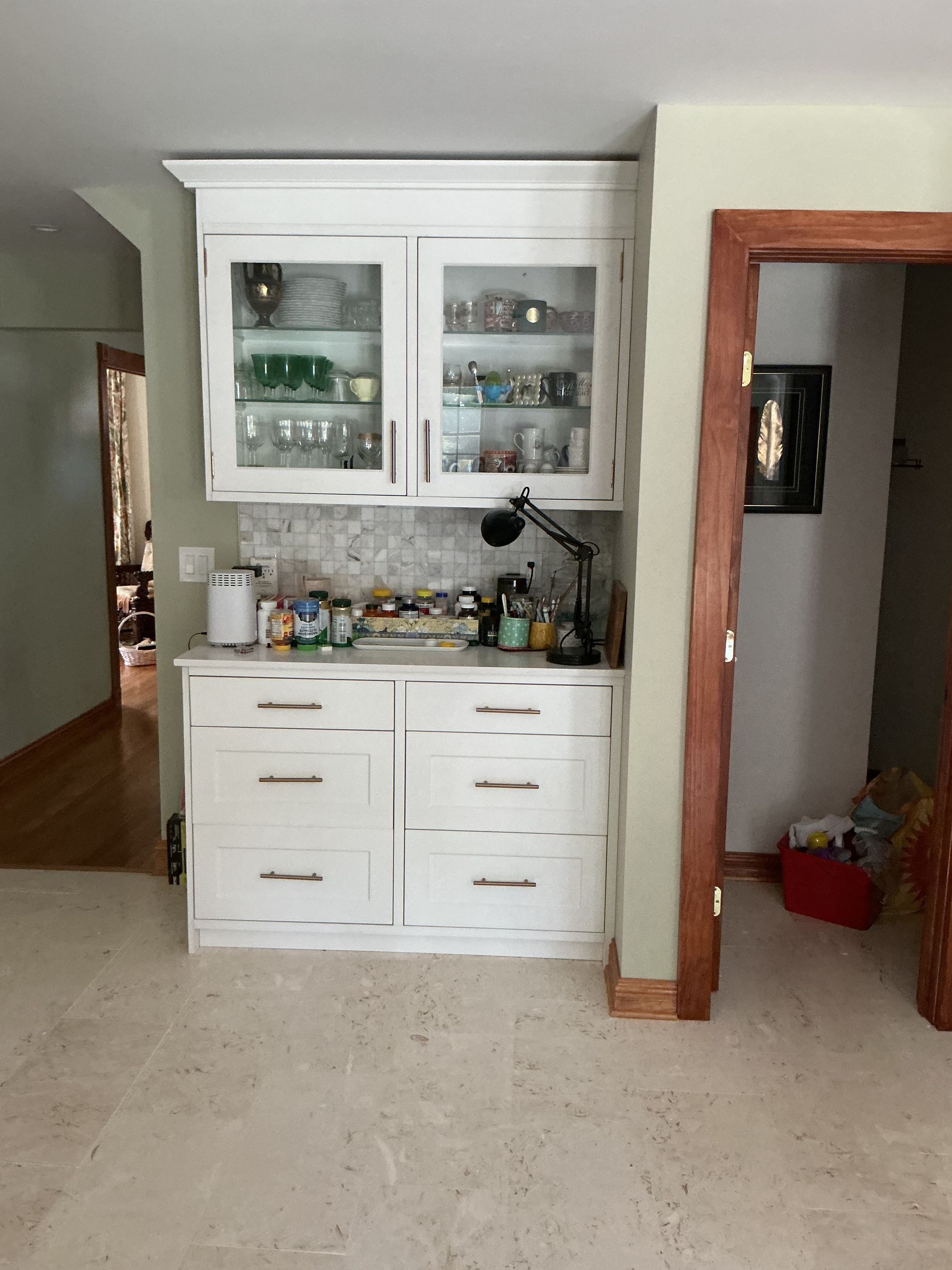 White kitchen hutch with glass cabinets and drawers in a beige room, with a doorway on the right.