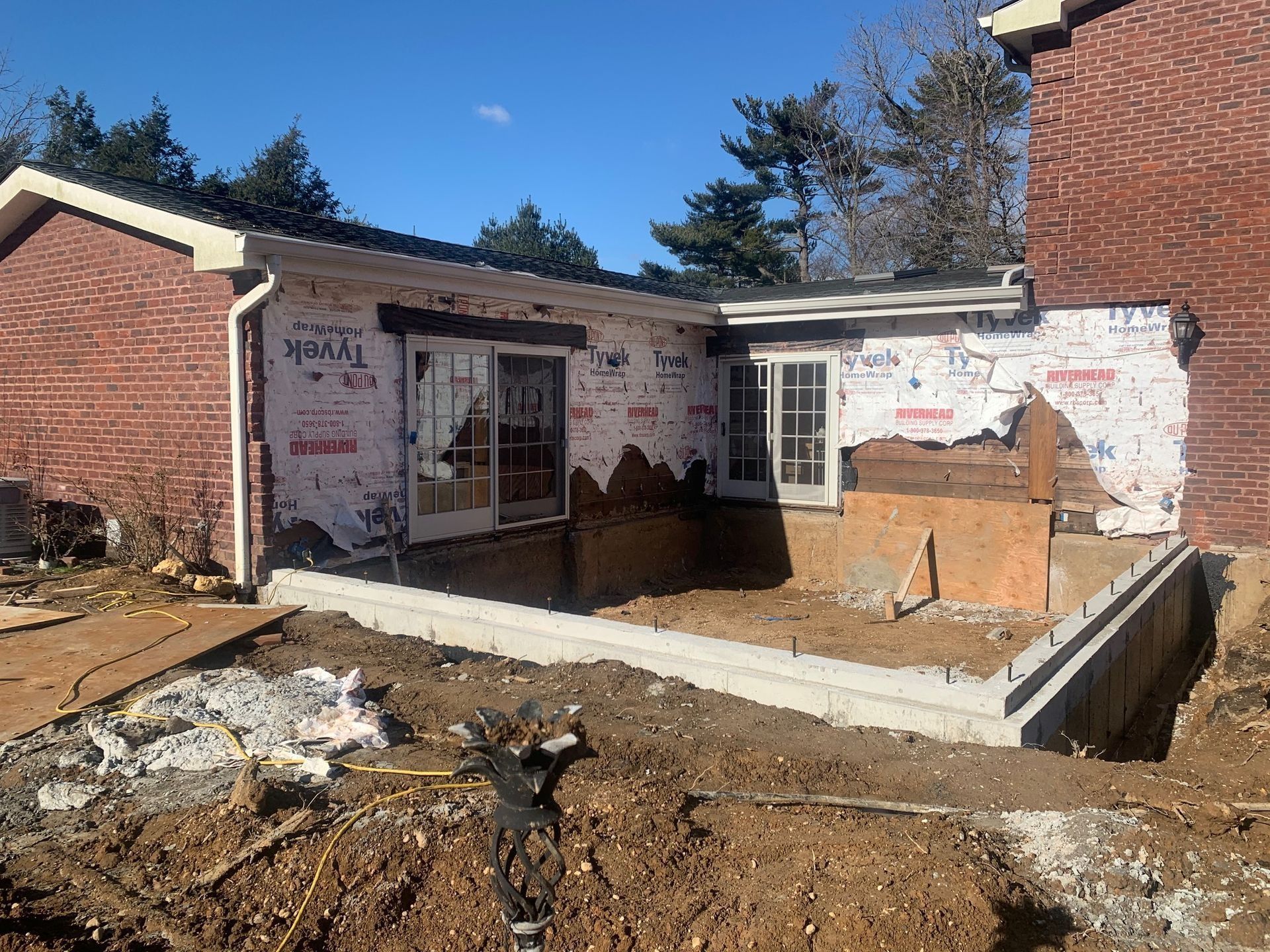 Construction site: building with exposed framing, Tyvek wrap, and new concrete foundation. Red brick walls and a clear sky.