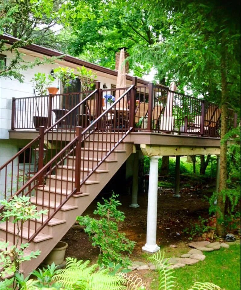 Elevated wooden deck with stairs and brown railings, supported by white pillars, surrounded by trees.