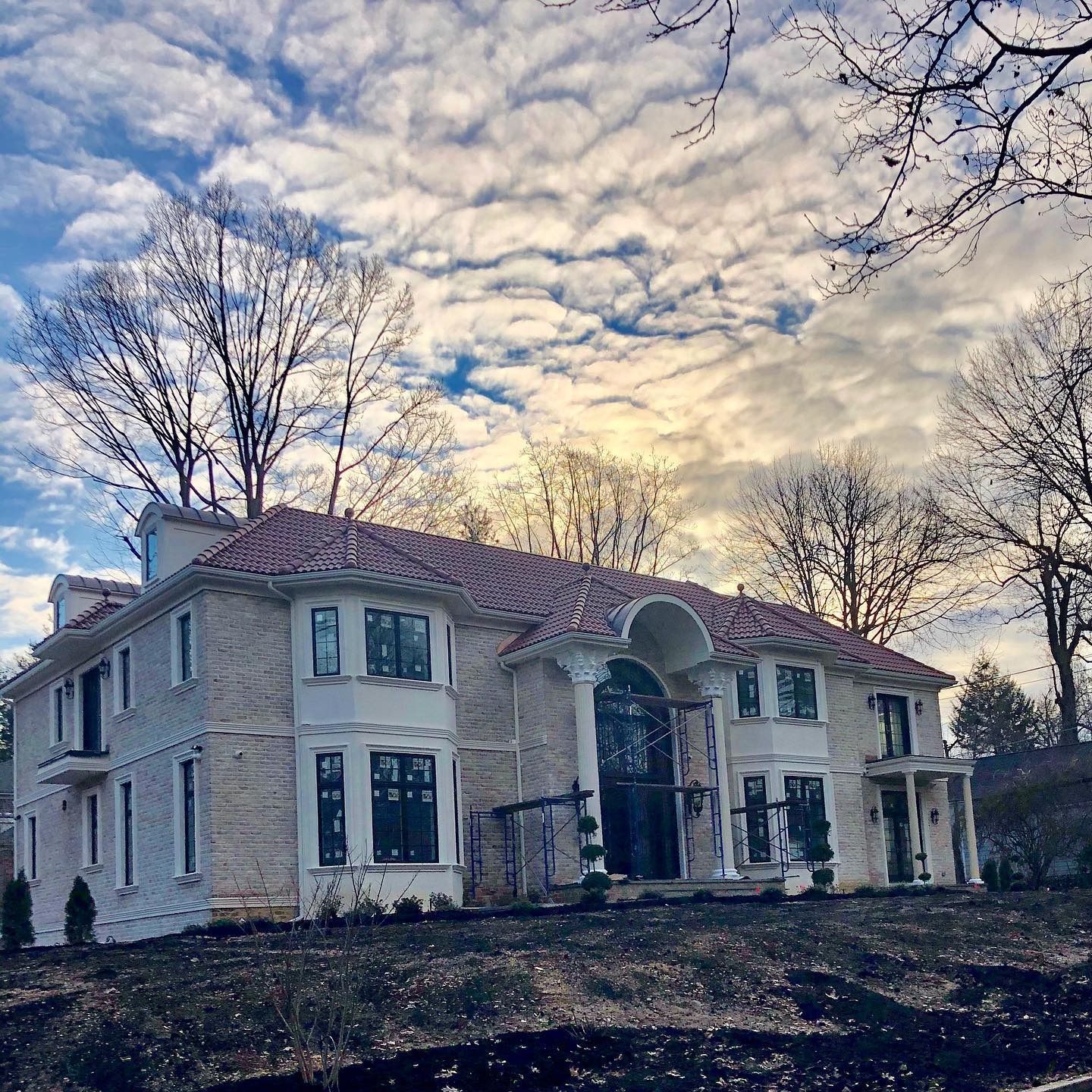 Two-story beige house with a red tile roof; bare trees and a cloudy sky in the background.
