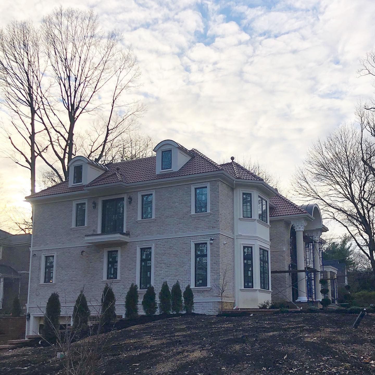 Two-story house with a red-tiled roof, light brick exterior, and columns, under a cloudy sky.