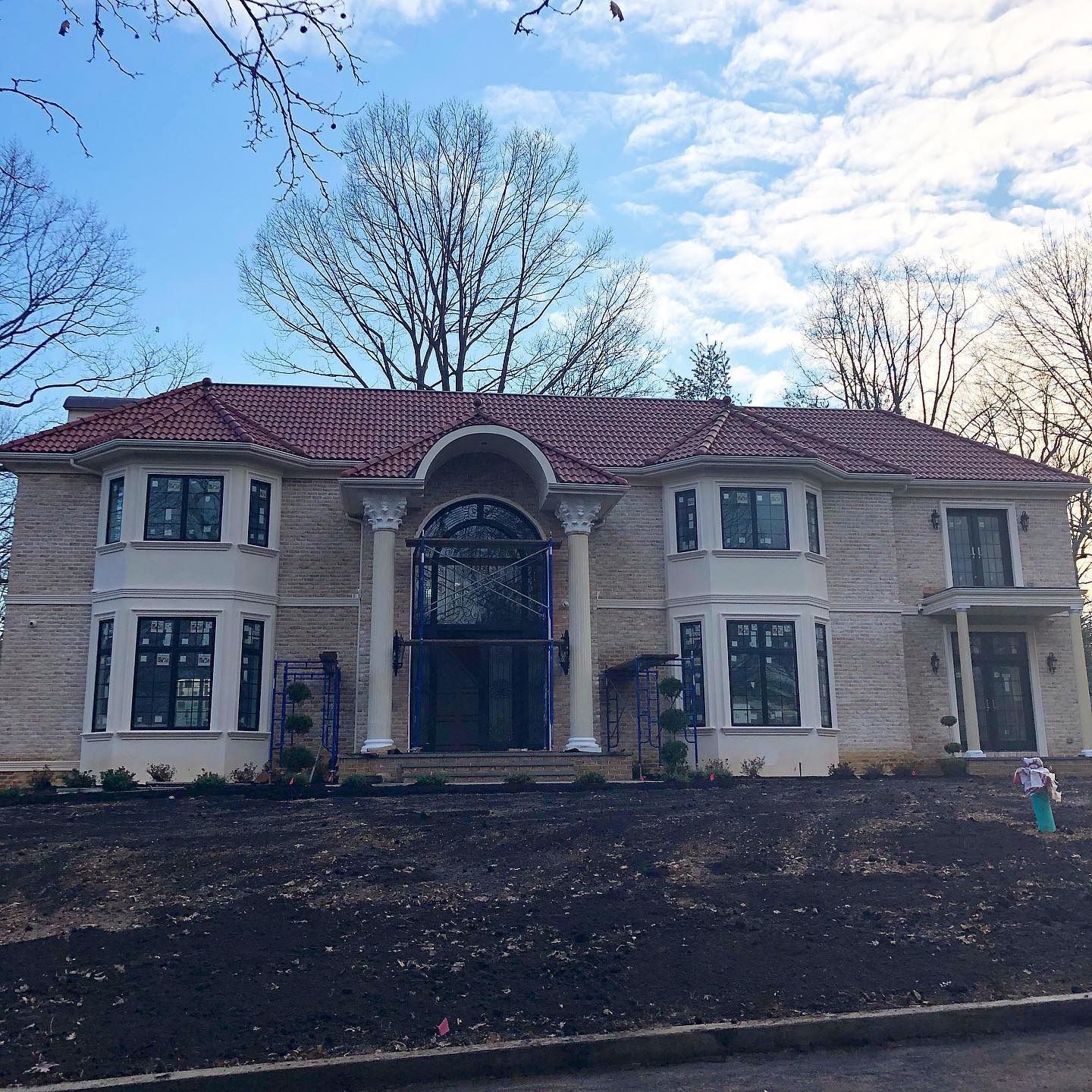 Large beige two-story house with red tile roof, pillars, and dark framed windows against a blue sky.