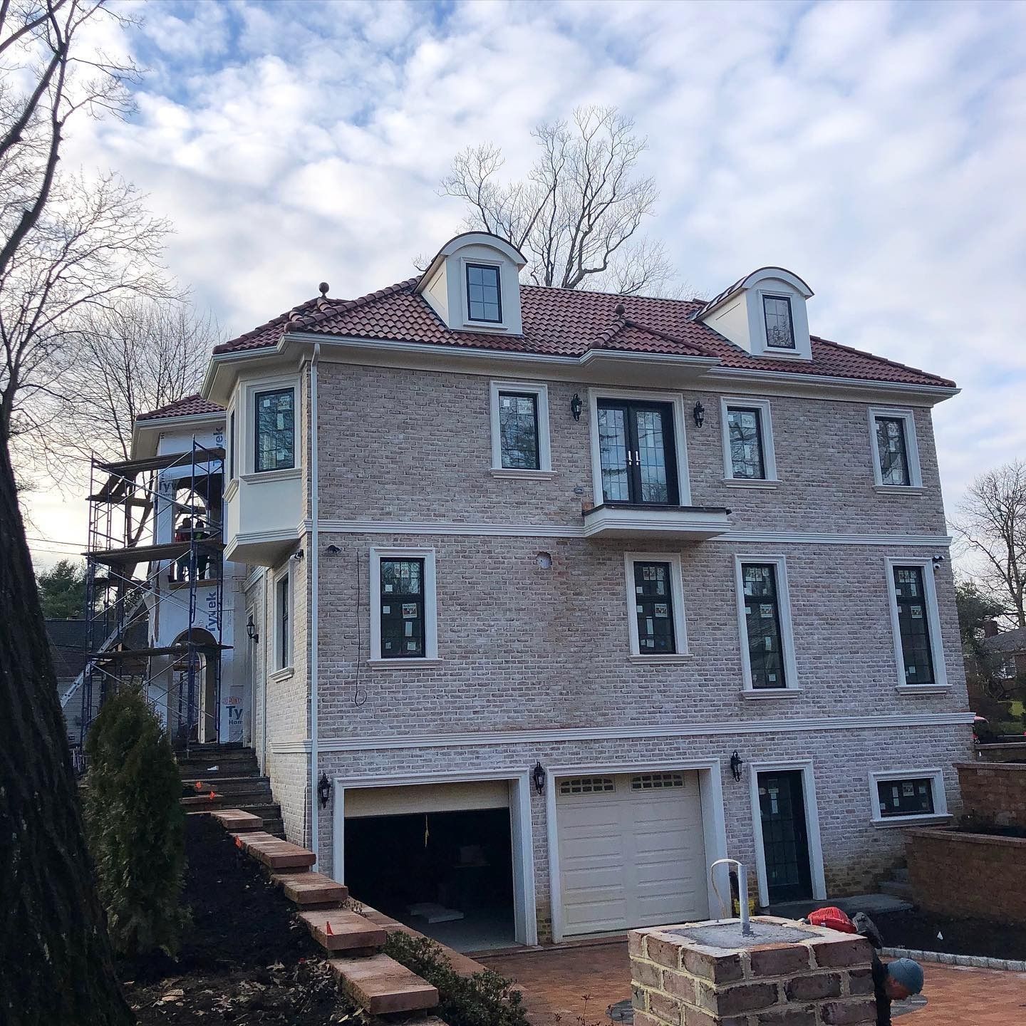 Three-story house with brick facade, red-tiled roof, dormers, and a two-car garage under a cloudy sky.