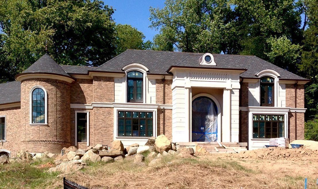 A house under construction with brick facade and gray roof, in a clearing, with trees in the background.