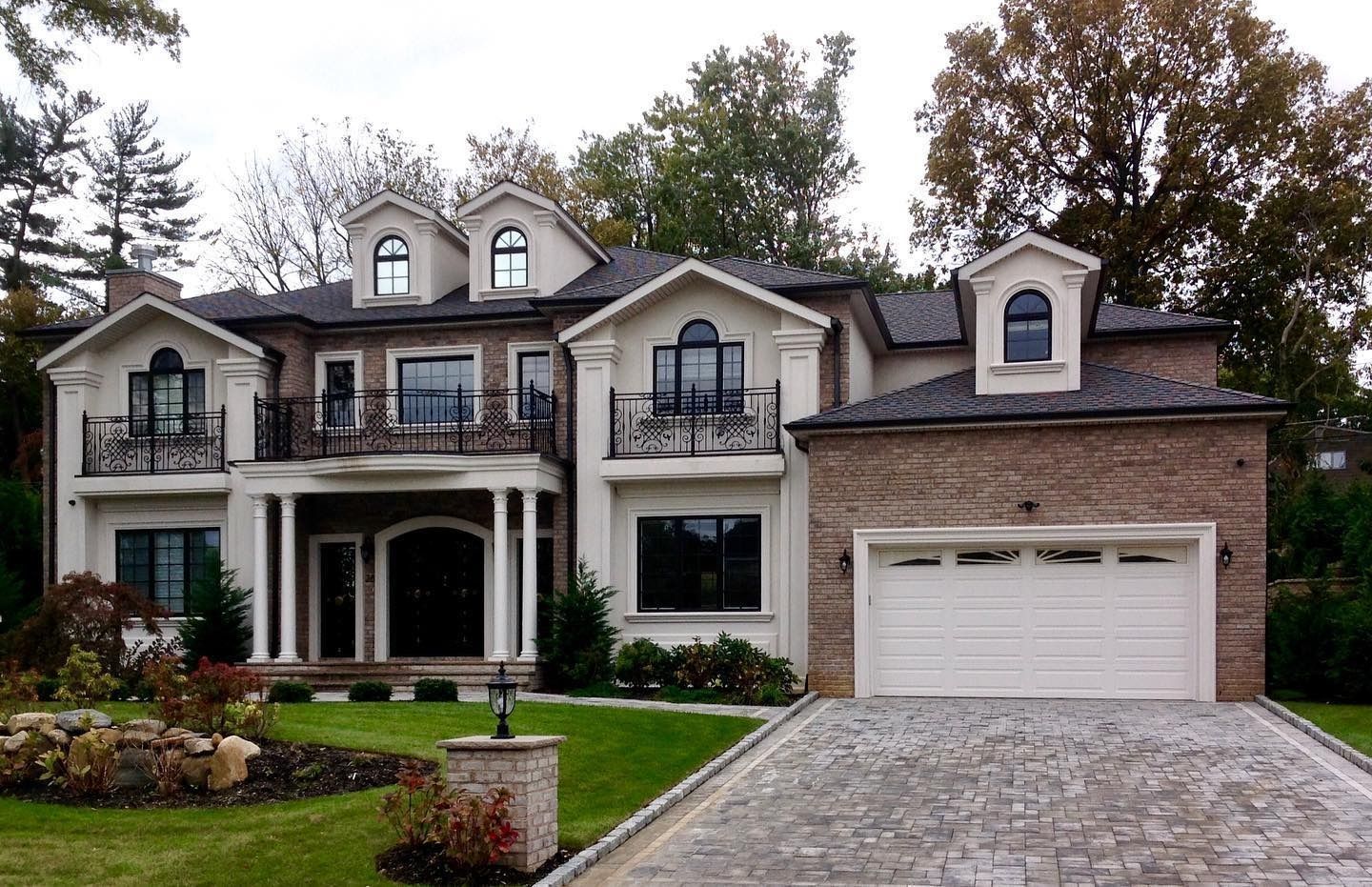 Two-story house with beige stucco, brick, and dark window frames. Features a garage and driveway.