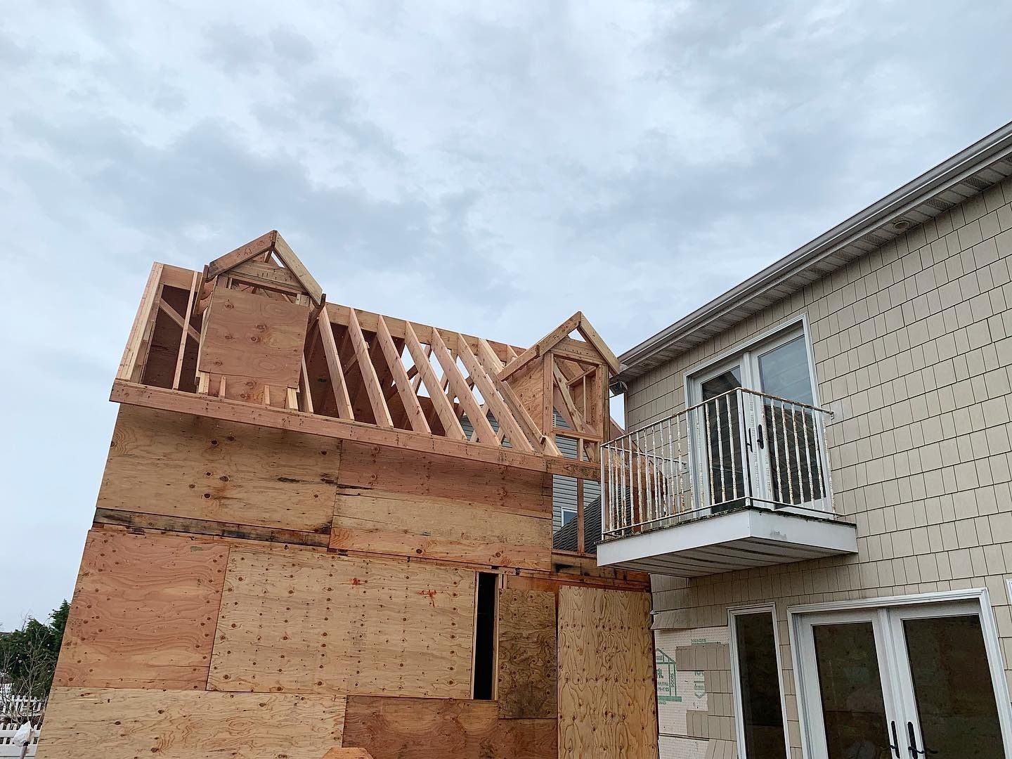 House under construction with exposed wood framing and a finished section with a balcony. Cloudy sky.